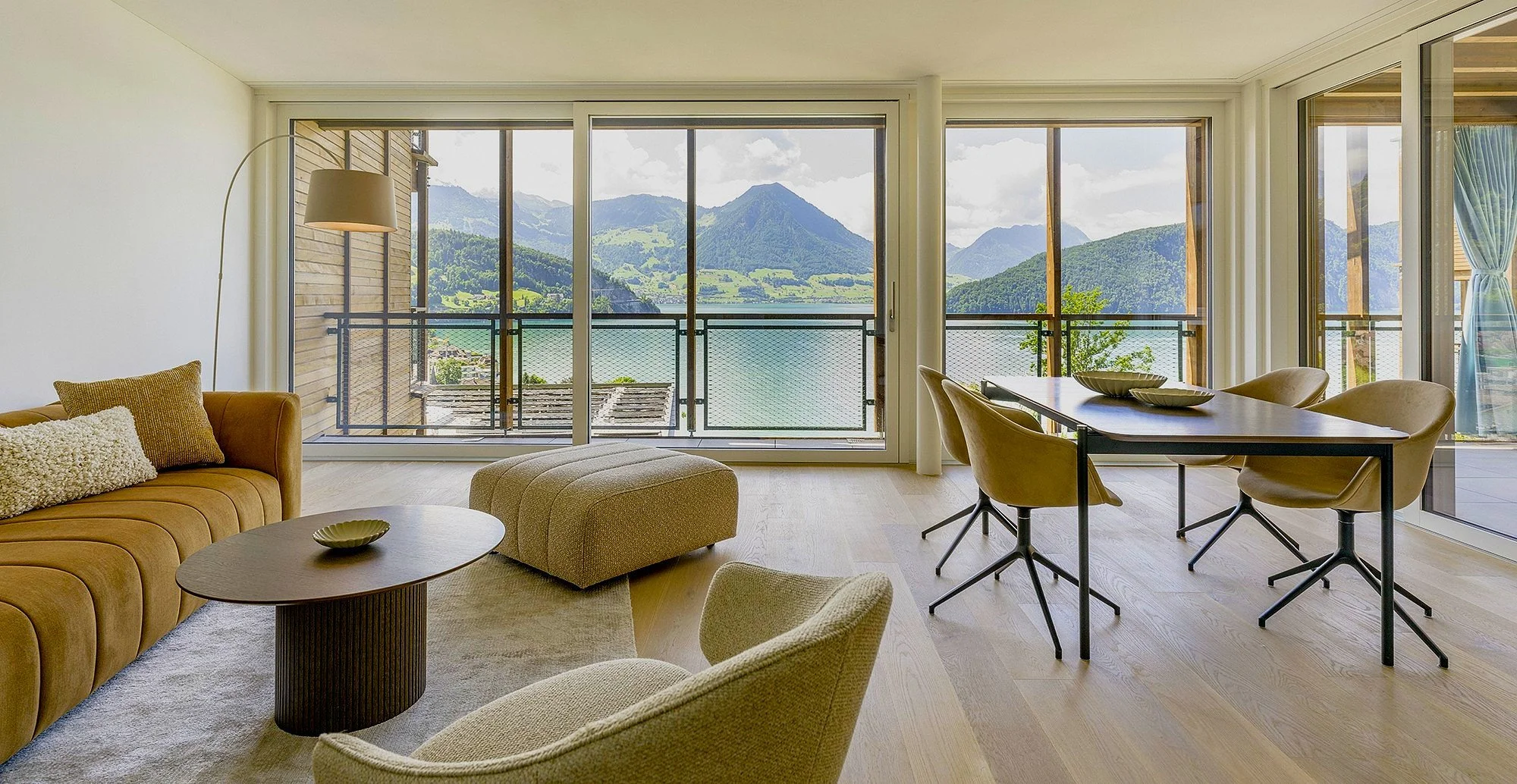 Wohnzimmer mit großen Fenstern und Blick auf Berge und Wasser, braunes Sofa, beige Sessel, Kaffeetisch, Esstisch mit Stühlen, moderne Einrichtung