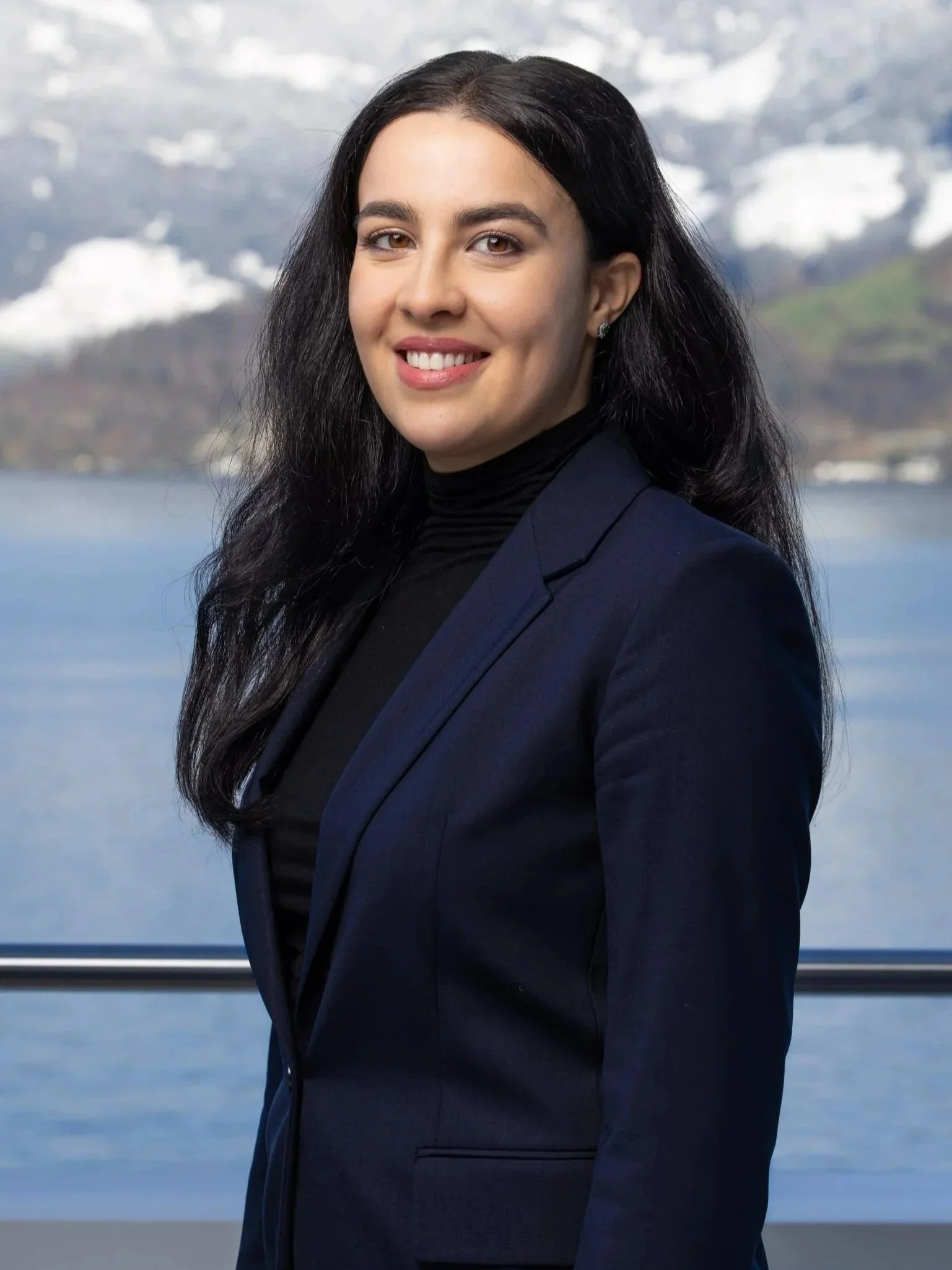 Junge Frau mit dunklen Haaren in dunklem Blazer vor einem See mit schneebedeckten Bergen im Hintergrund.
