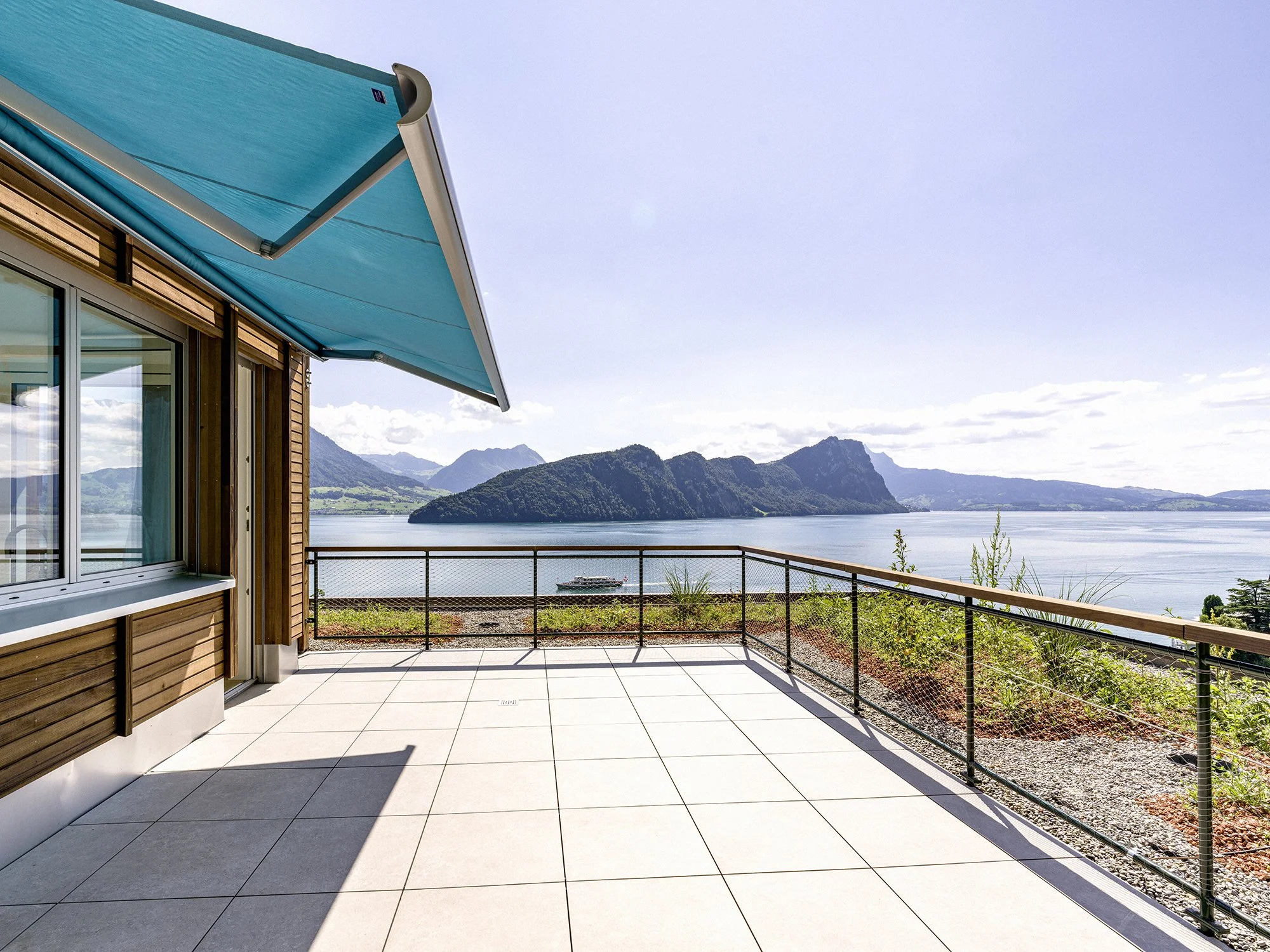 Balkon mit Blick auf den See, Berge im Hintergrund, blauer Himmel, Sonnenschutz im Überhang, Glasfenster und Holzfassade