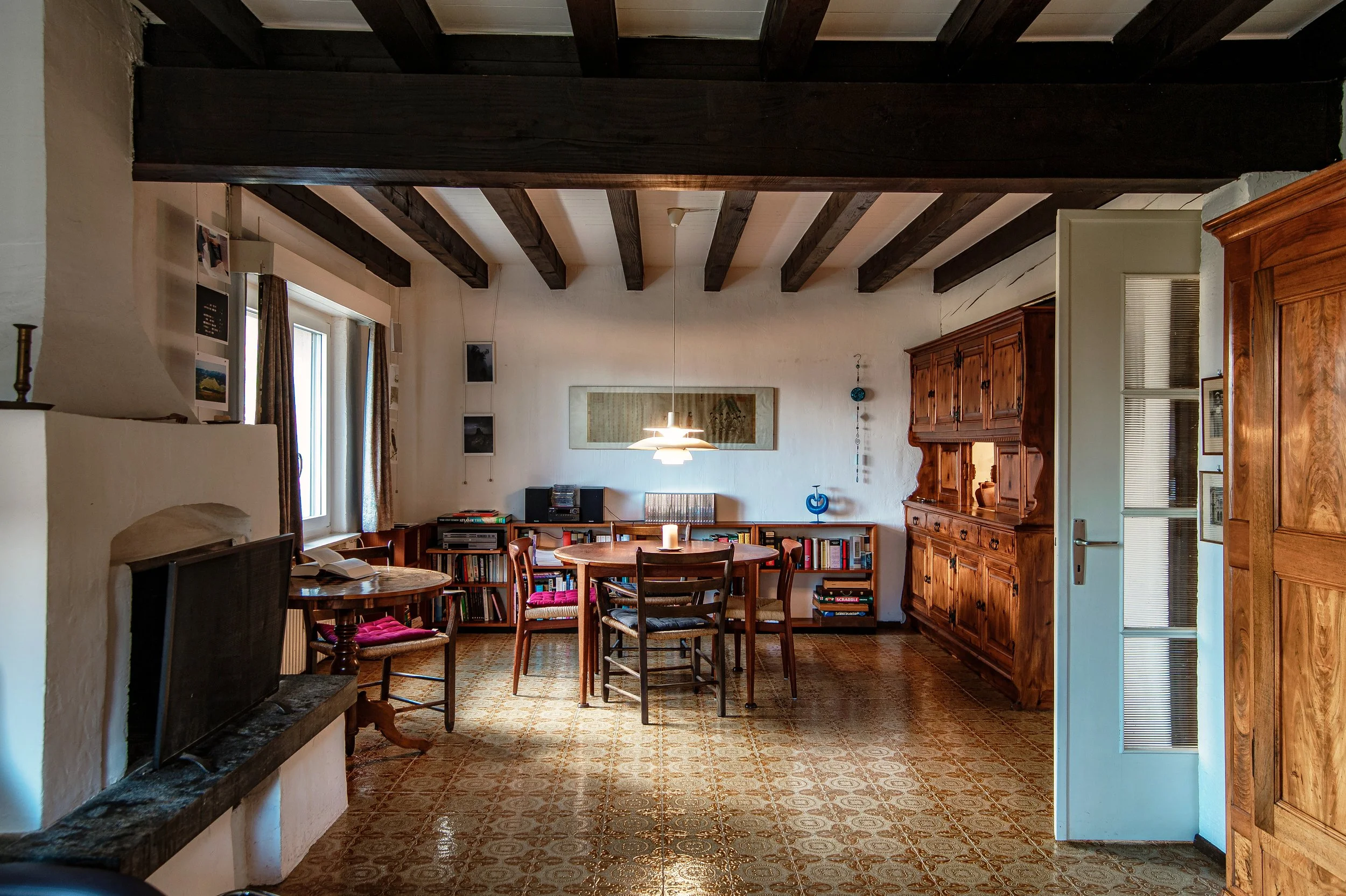 Gemütliches Wohnzimmer mit Holzbalken an der Decke, rustikaler Holzschrank, Esstisch mit Stühlen, Bücherregale, Wandbilder und eine Lampe.