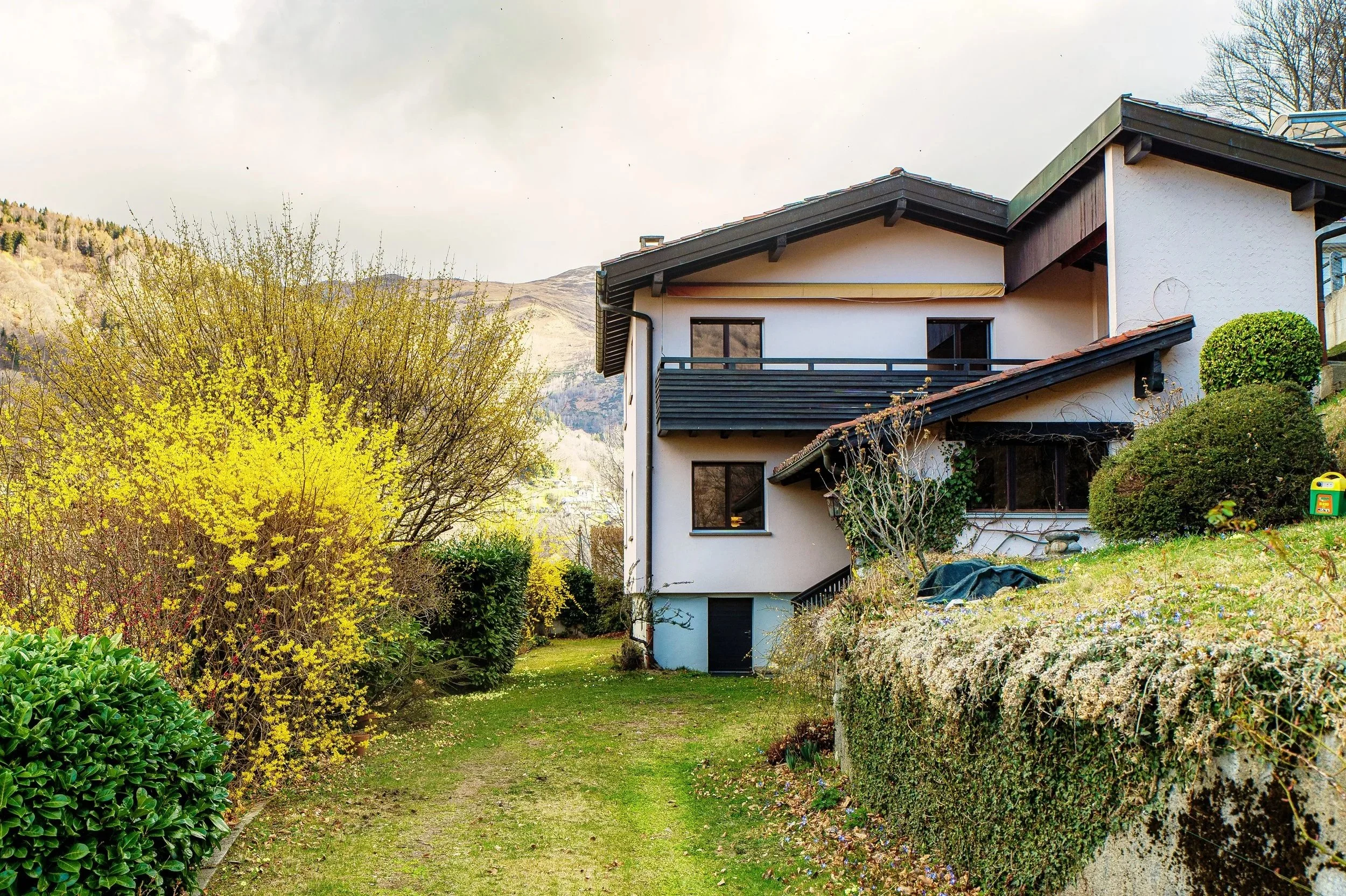 Mehrstöckiges Haus mit Balkon, umgeben von Büschen und Bäumen auf einer Hügellandschaft im Frühling.