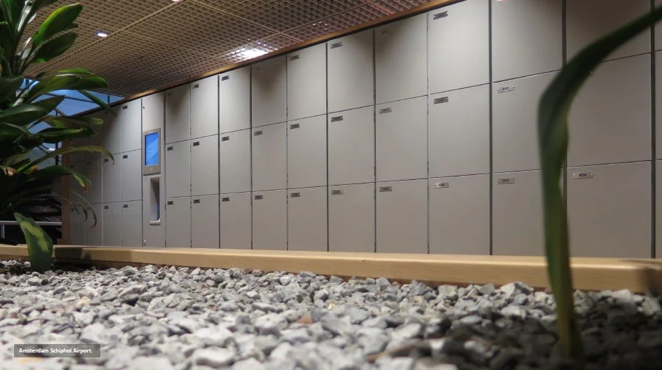 Airport storage lockers against a wall with a gravel floor and some plants in the foreground.