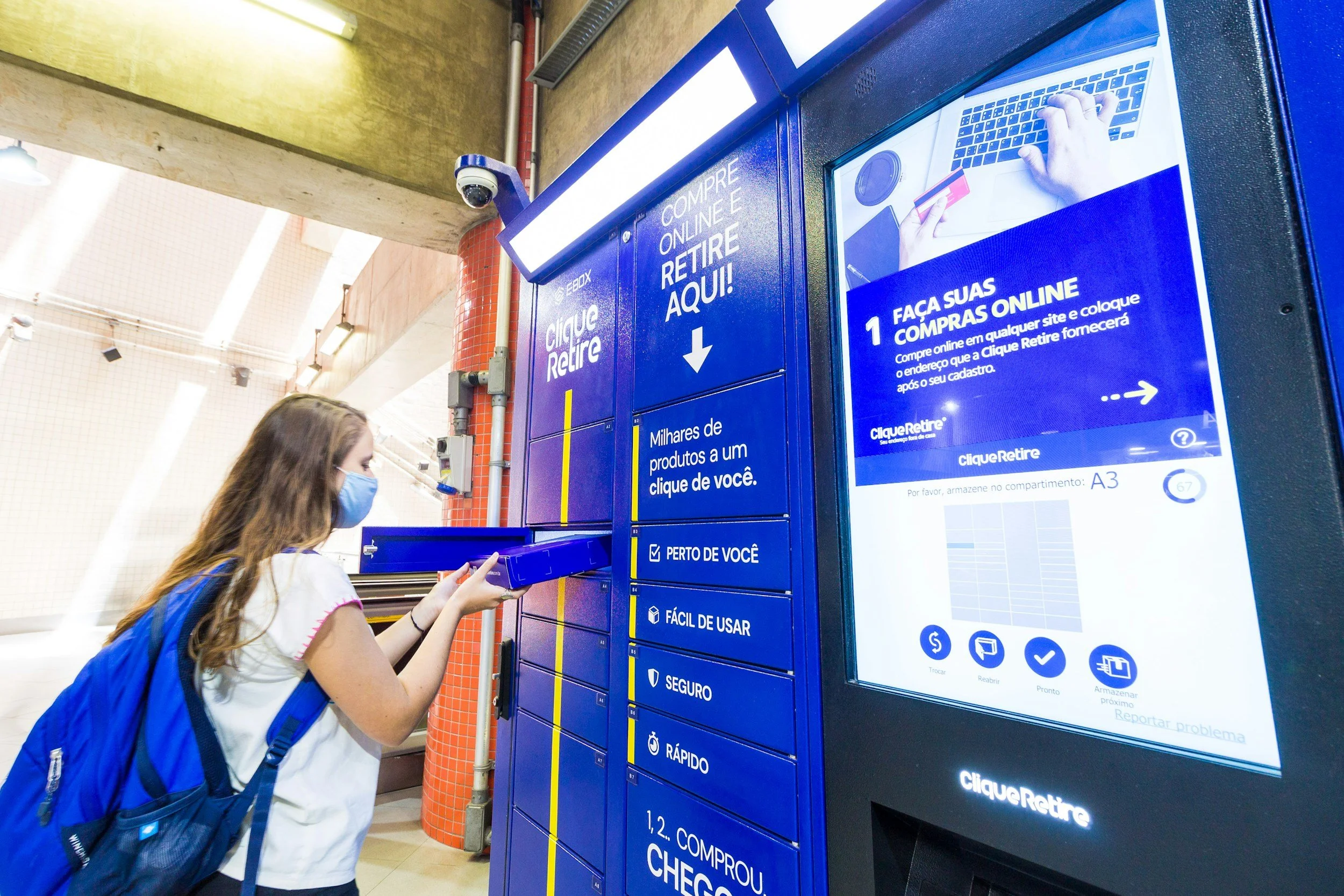 A young woman wearing a face mask and a backpack using a parcel locker to pick up an online purchase at a parcel pickup station.