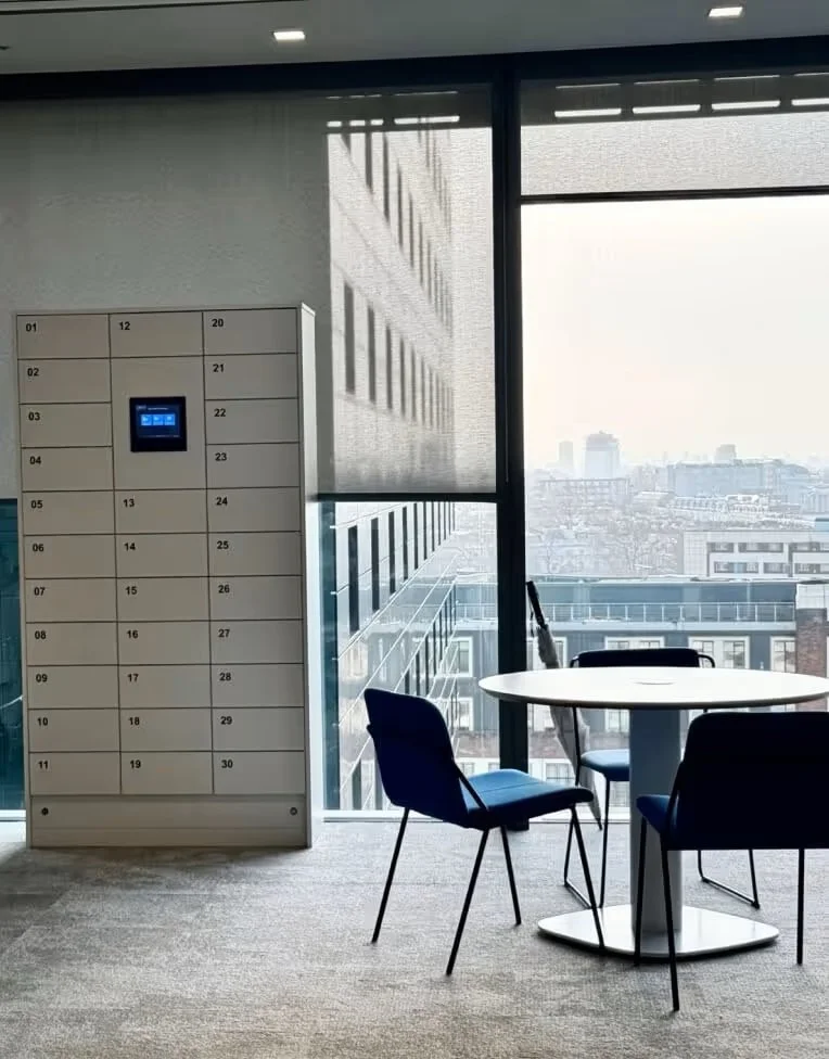 An office lounge area with a round white table, three blue chairs, and large windows overlooking a city sky and buildings, with a modern locker or kiosk with a small screen.
