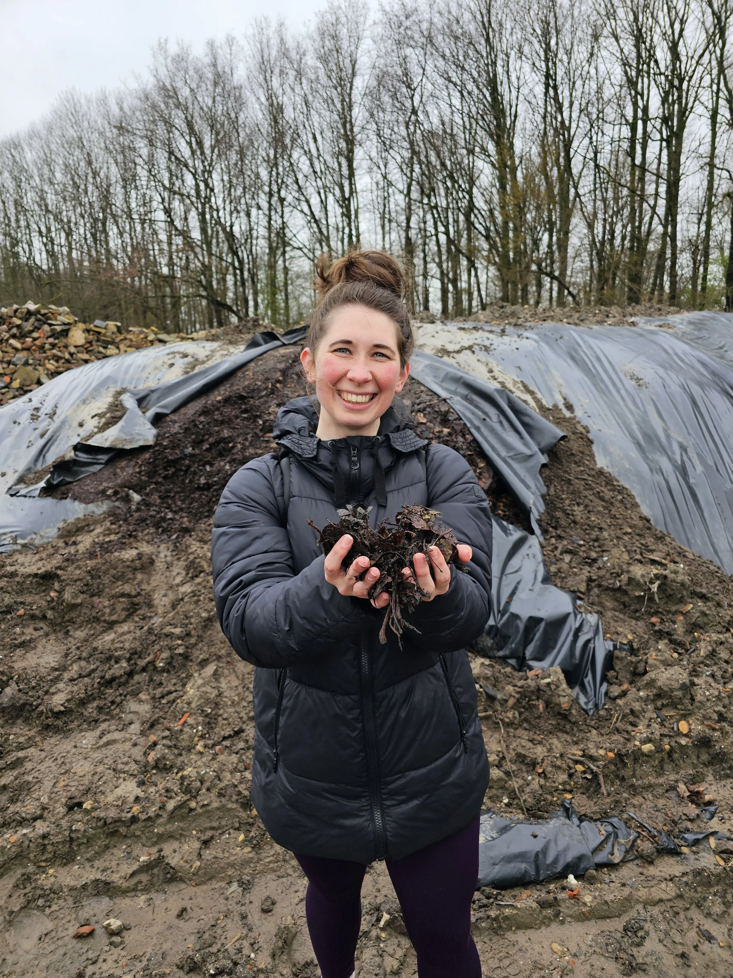 A woman in a black jacket holding a clump of soil or compost, standing in front of a large pile of soil covered with a black plastic sheet outdoors during the daytime, with leafless trees in the background.
