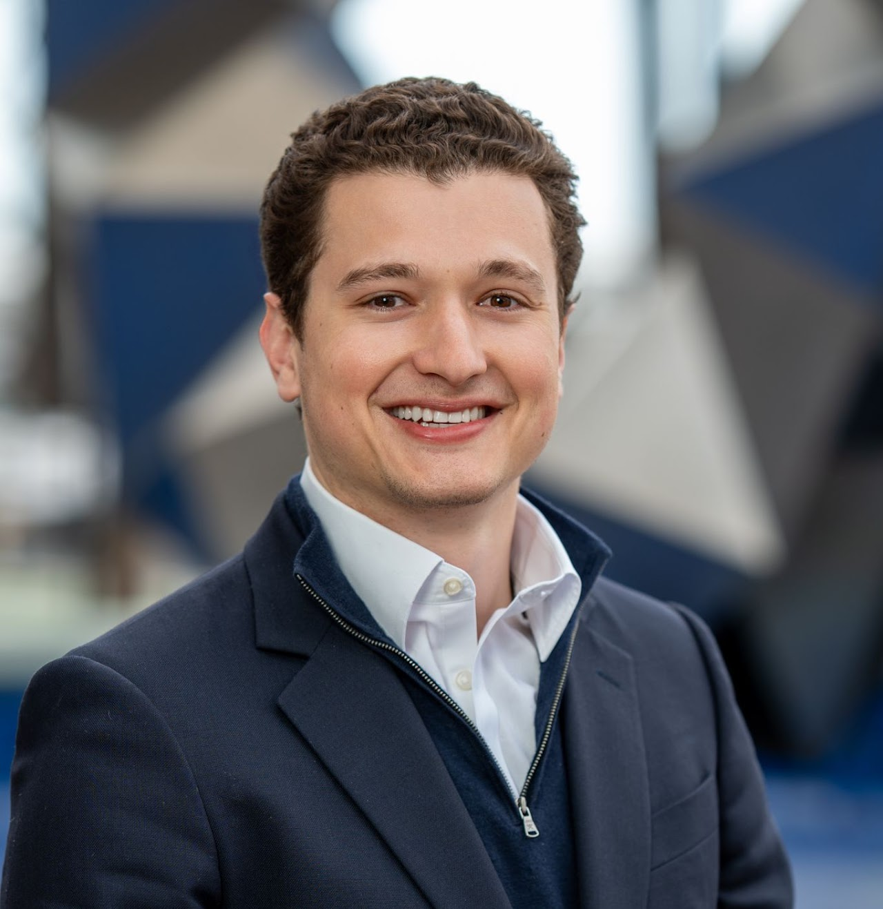 A smiling young man wearing a blazer and white shirt standing outdoors with blurred buildings and structures in the background.