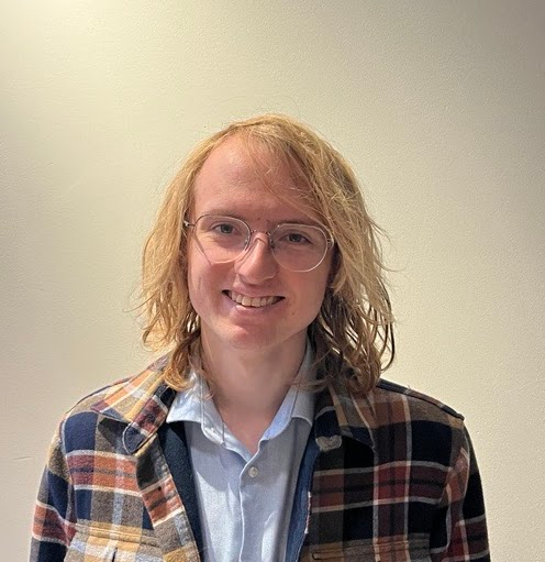 A young person with shoulder-length blond hair, glasses, and a light blue shirt, smiling at the camera, wearing a plaid jacket, standing against a plain light-colored wall.