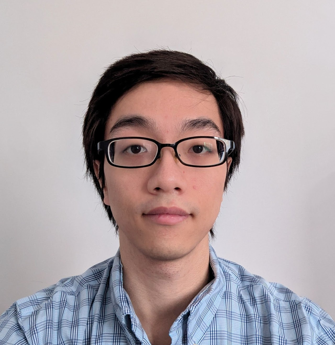 Portrait of a young man with glasses, wearing a blue checkered shirt, standing against a plain white background.