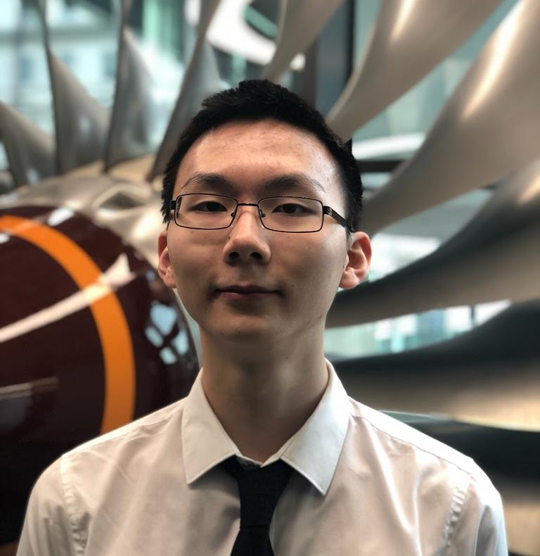 A young man wearing glasses, a white shirt, and a black tie, smiling softly, standing in front of a modern, metallic architectural structure with a large, spherical object in the background.