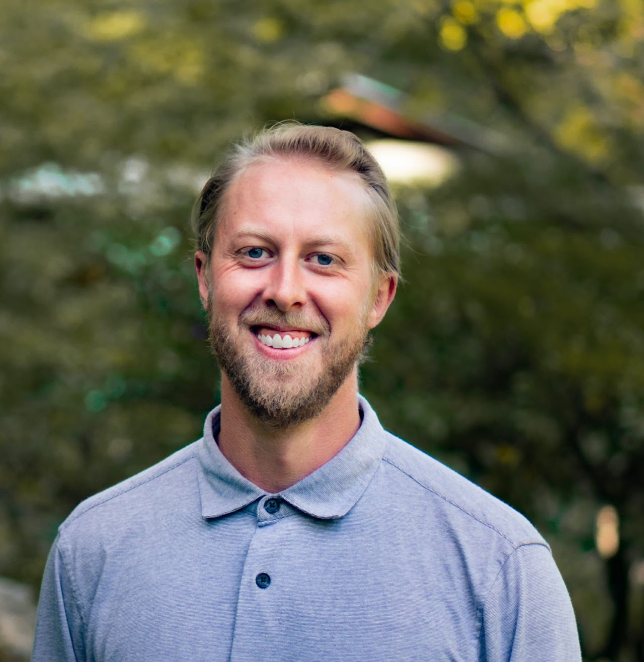 A smiling man with a beard and blue eyes wearing a light gray polo shirt, standing outdoors with blurred green trees in the background.