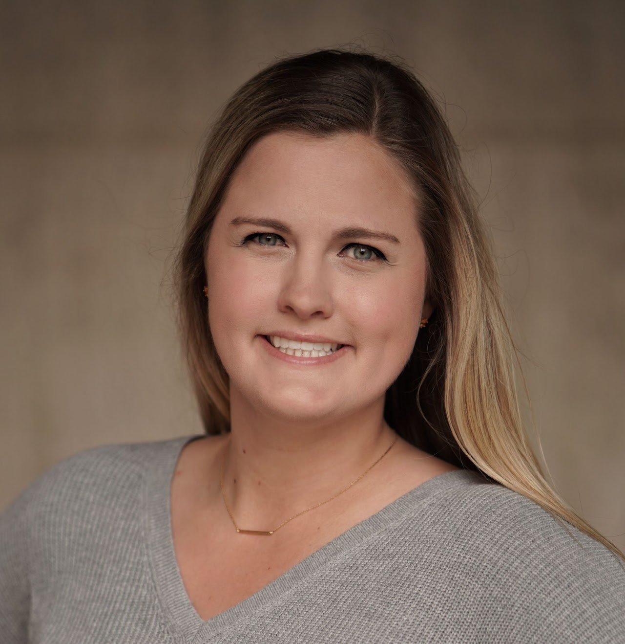 Headshot of a smiling woman with light brown hair, blue eyes, wearing a gray top and gold necklace, against a beige background.