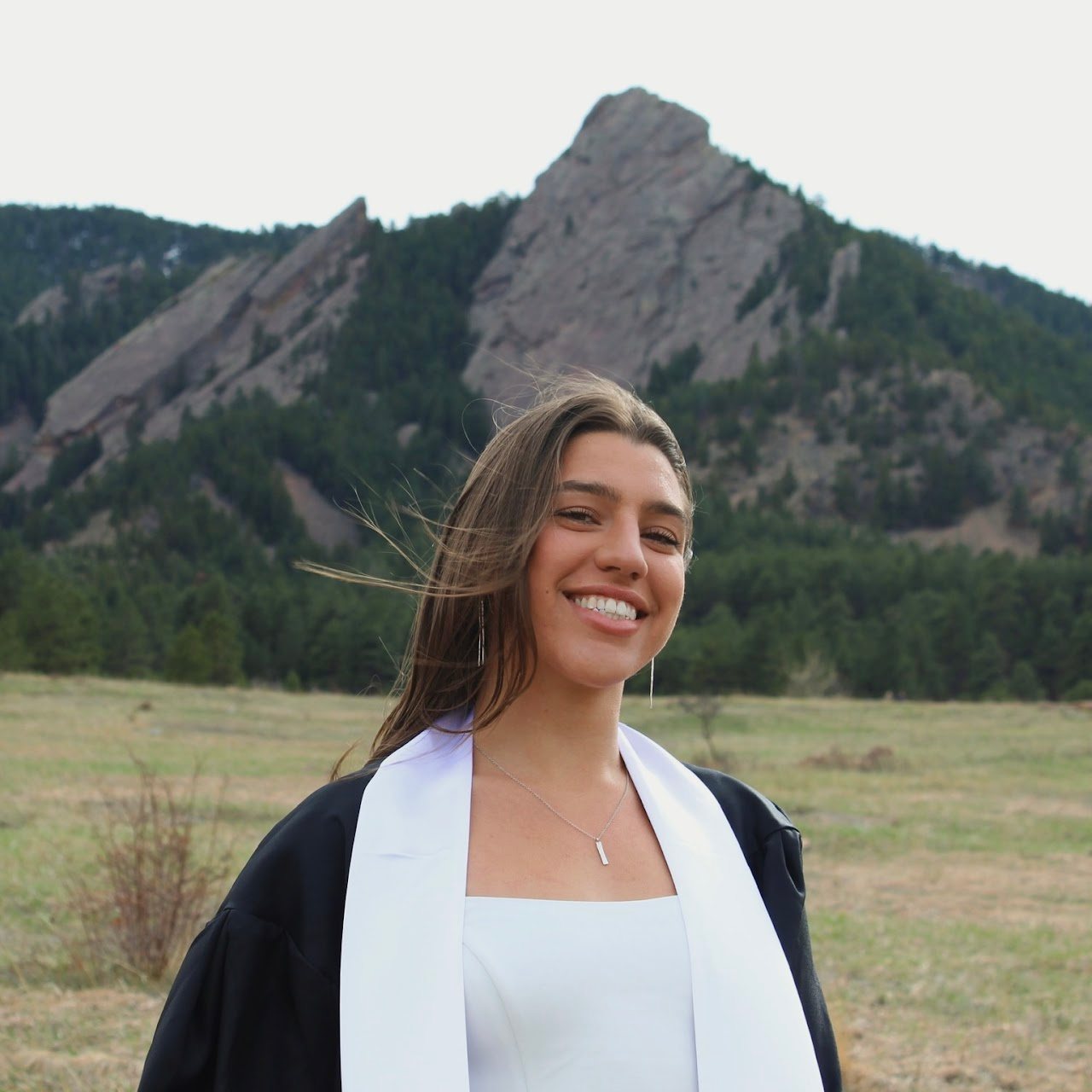 Young woman in graduation gown smiling outdoors with mountains and forest in the background.
