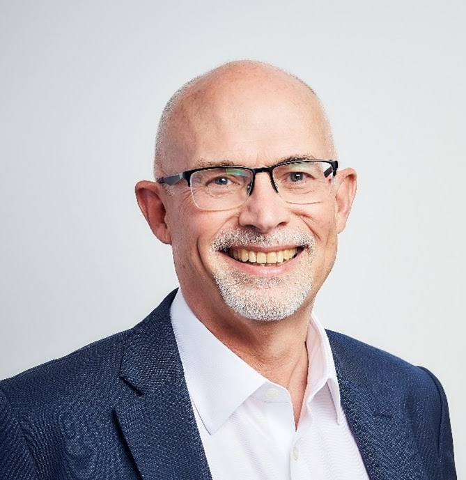 A smiling man with glasses, a white beard, wearing a blue blazer and a white shirt, posing against a plain light background.