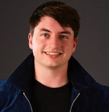 A young man with short brown hair and fair skin smiling, wearing a dark jacket over a black shirt, against a dark background.