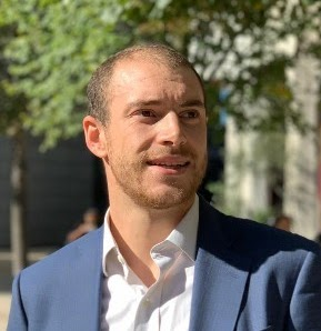 A man in a blue suit and white shirt standing outdoors with trees in the background.