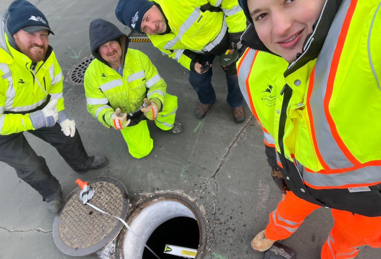 Four workers in high-visibility jackets and safety gear are gathered around an open manhole, inspecting and taking notes at a street repair site.
