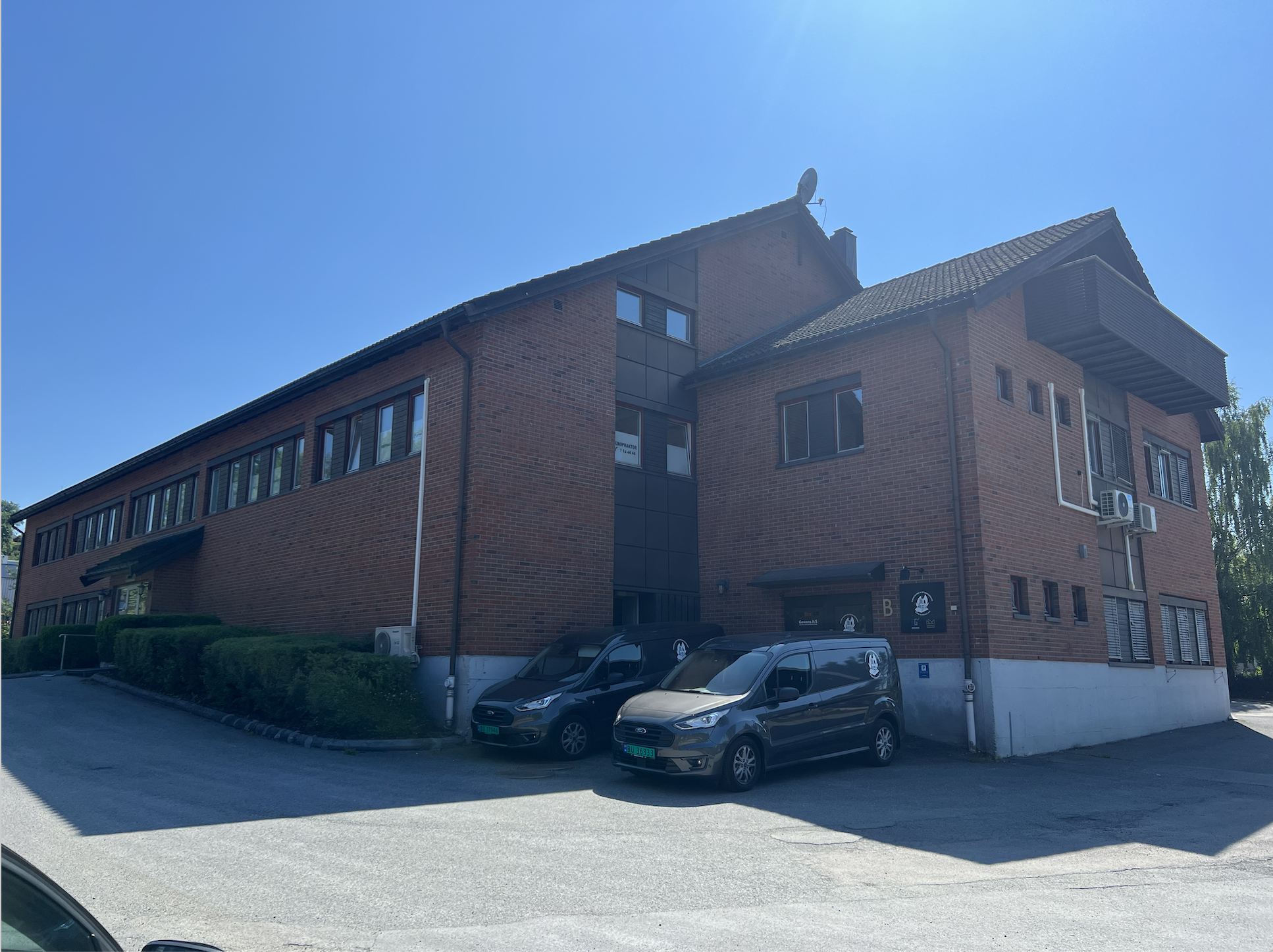 A multi-story brick building with several windows, air conditioning units, and parked cars in front under a clear blue sky.