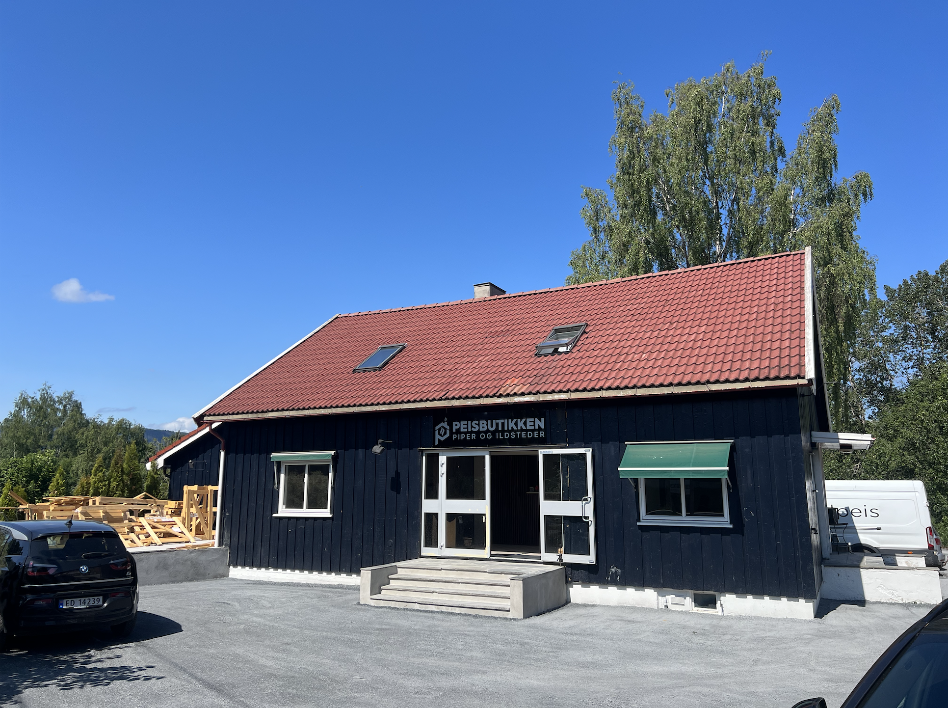 A black wooden building with a red tile roof and two skylights, exterior with two windows and a glass door with open double doors, steps leading up to the entrance, a sign reading 'PEISBUTIKKEN PIPER OG ILDSTEDER', parking lot with cars and surrounding trees under a blue sky.