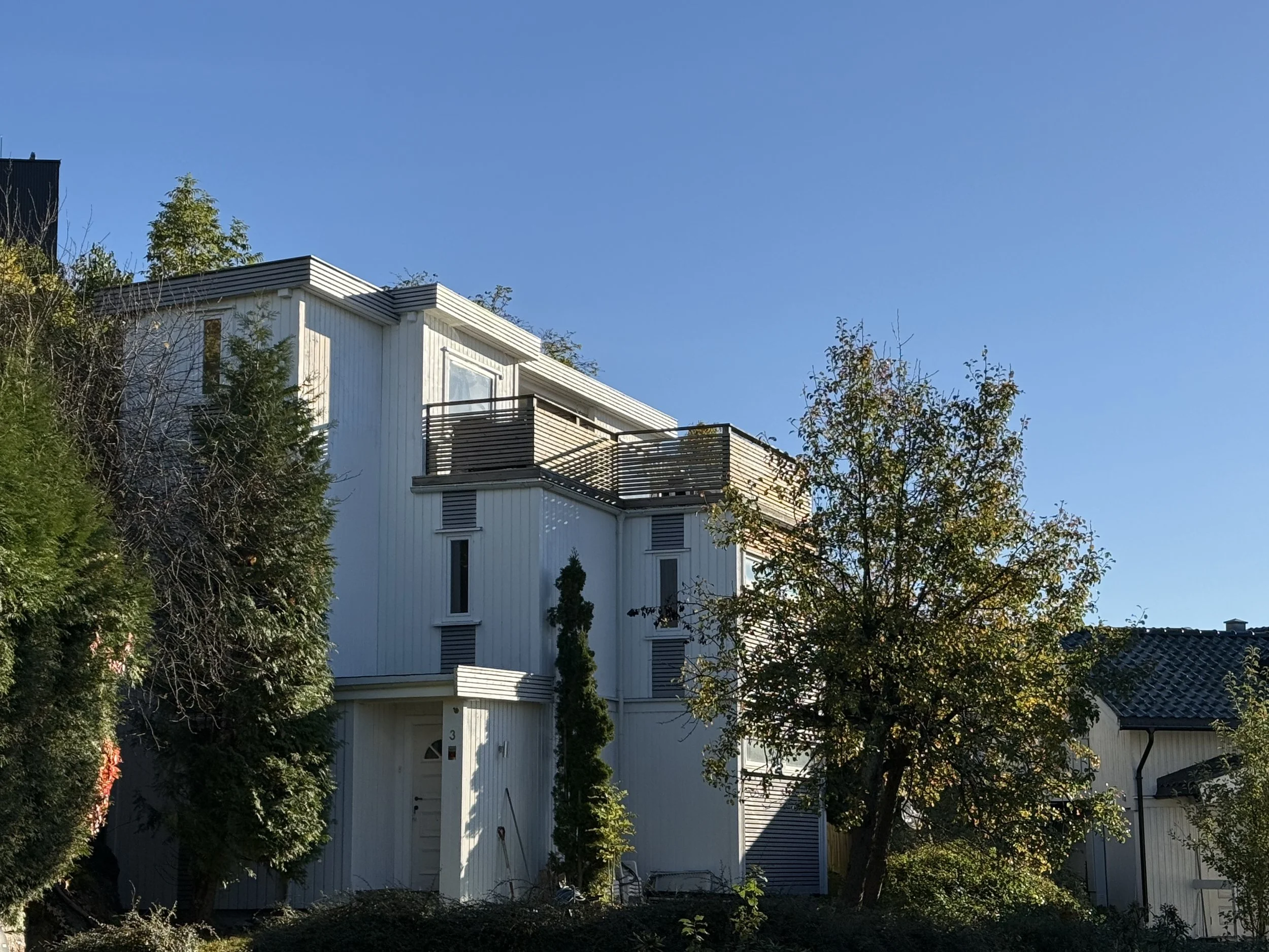 A modern white multi-story house with a balcony, surrounded by trees and bushes on a clear, sunny day.