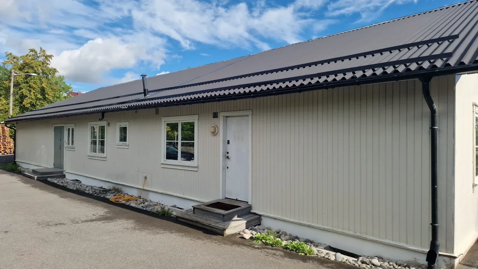 A long, white, single-story building with a gray metal roof, three small windows, and a white door with steps, situated beside a pavement with rocks and some plants, under a partly cloudy blue sky.