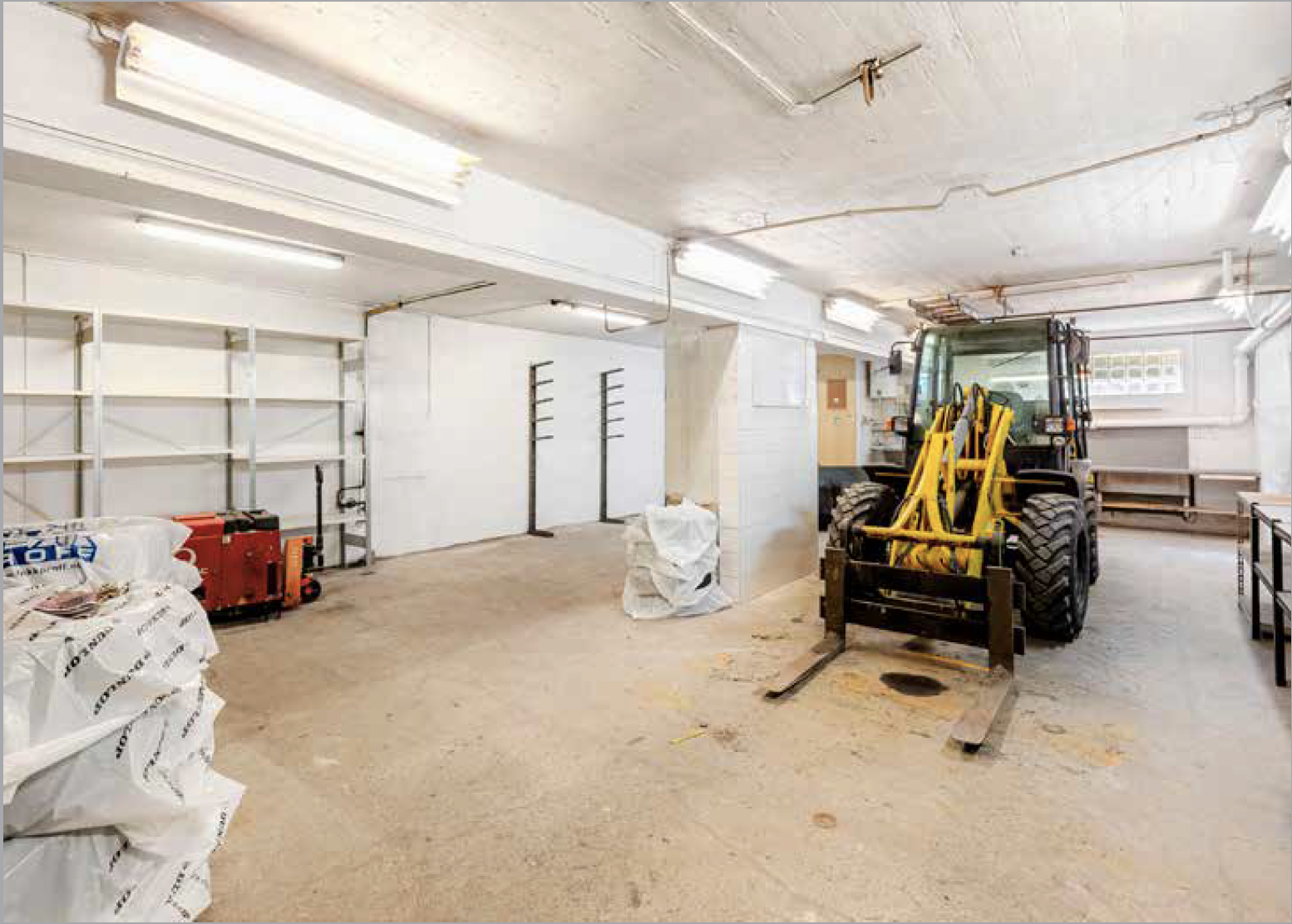 Empty garage or workshop space with white walls and ceiling, fluorescent lighting, a forklift, a red portable heater, and several bags or sacks on the floor.