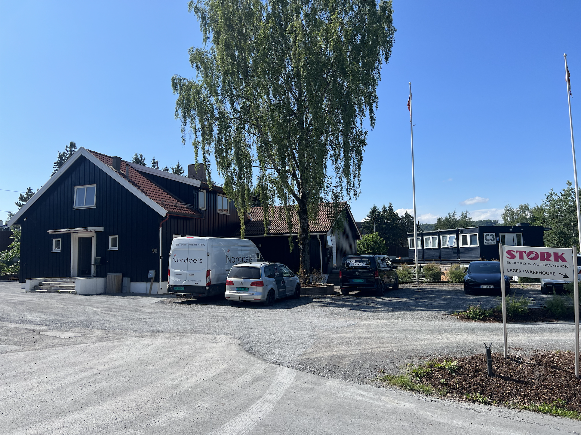 Parking lot with several parked cars near a black building, a large tree, and a sign for a warehouse store called Stork, with two flagpoles, under a blue sky with some clouds.