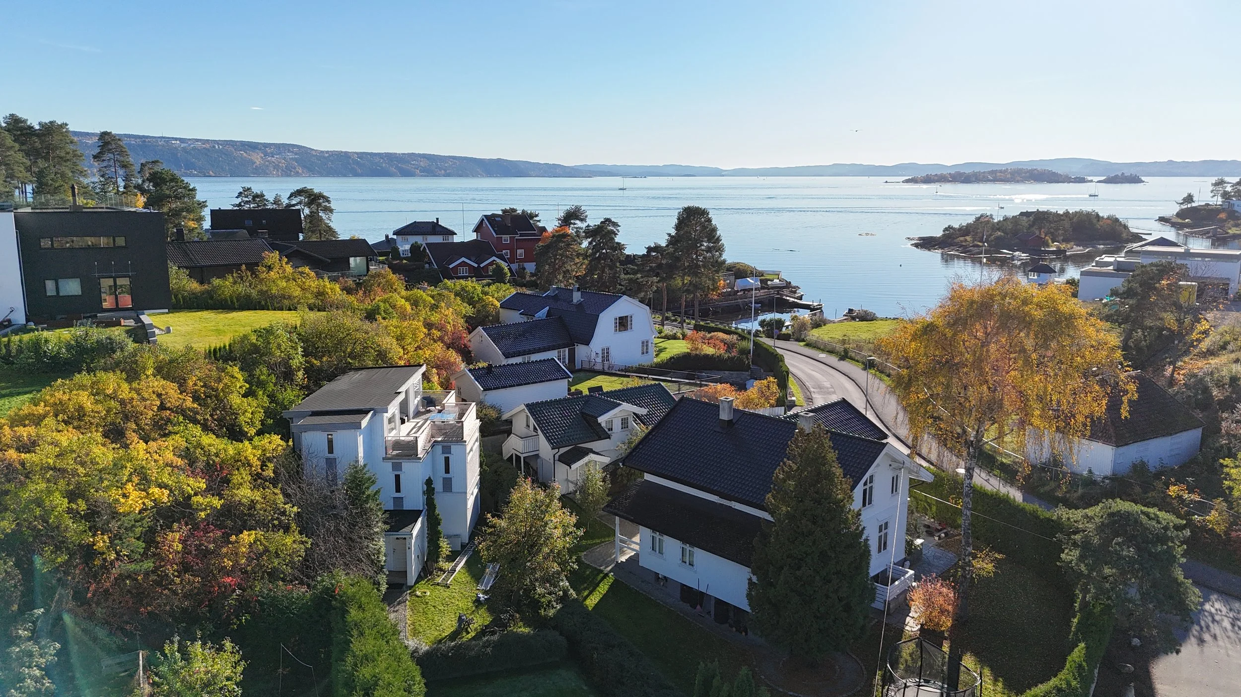 Aerial view of a scenic coastal neighborhood with white houses and lush trees near a calm body of water, under a clear blue sky.