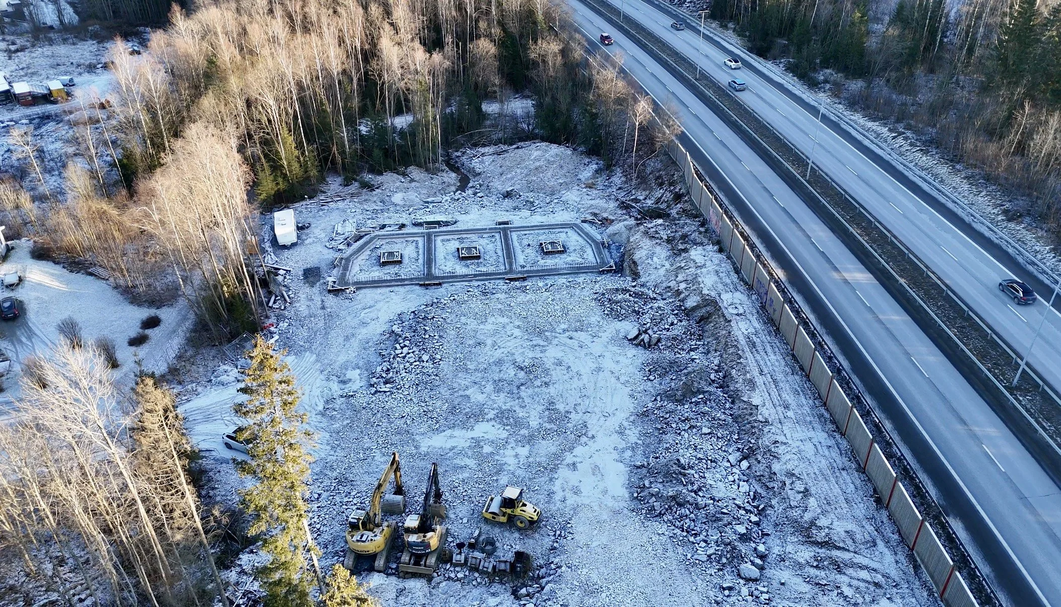 A construction site surrounded by snow, with excavators, construction materials, and a foundation work proceeding, adjacent to a highway with moving vehicles and a wooded area in winter.
