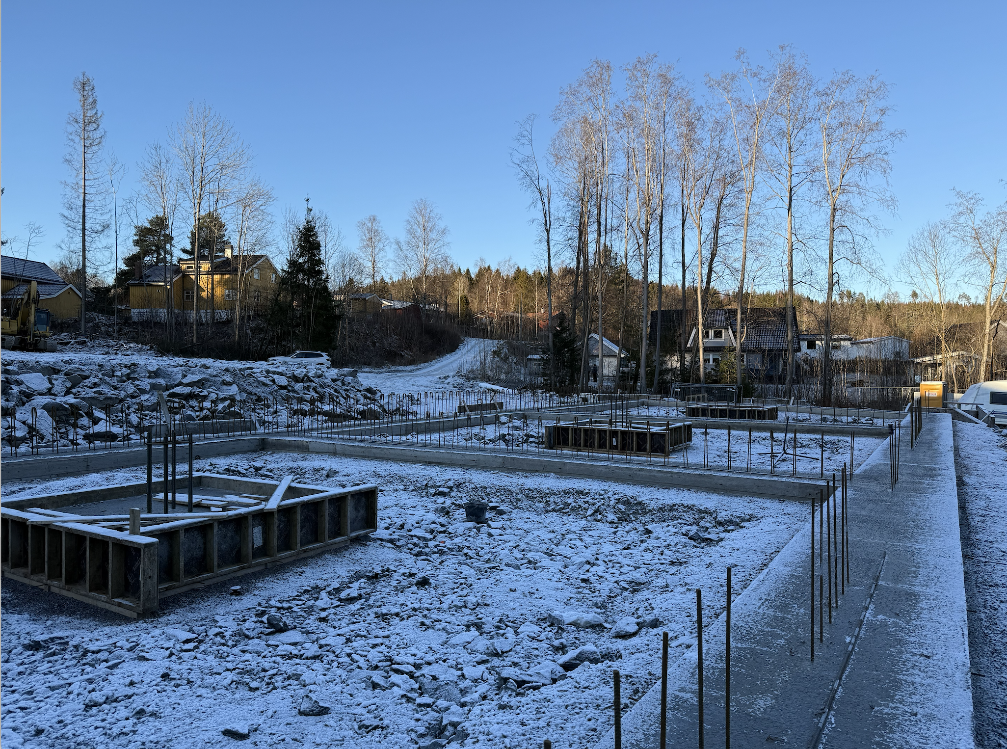 Construction site in a snowy landscape with foundation frames for building. There is a sidewalk on the right side and houses and trees in the background under a clear blue sky.