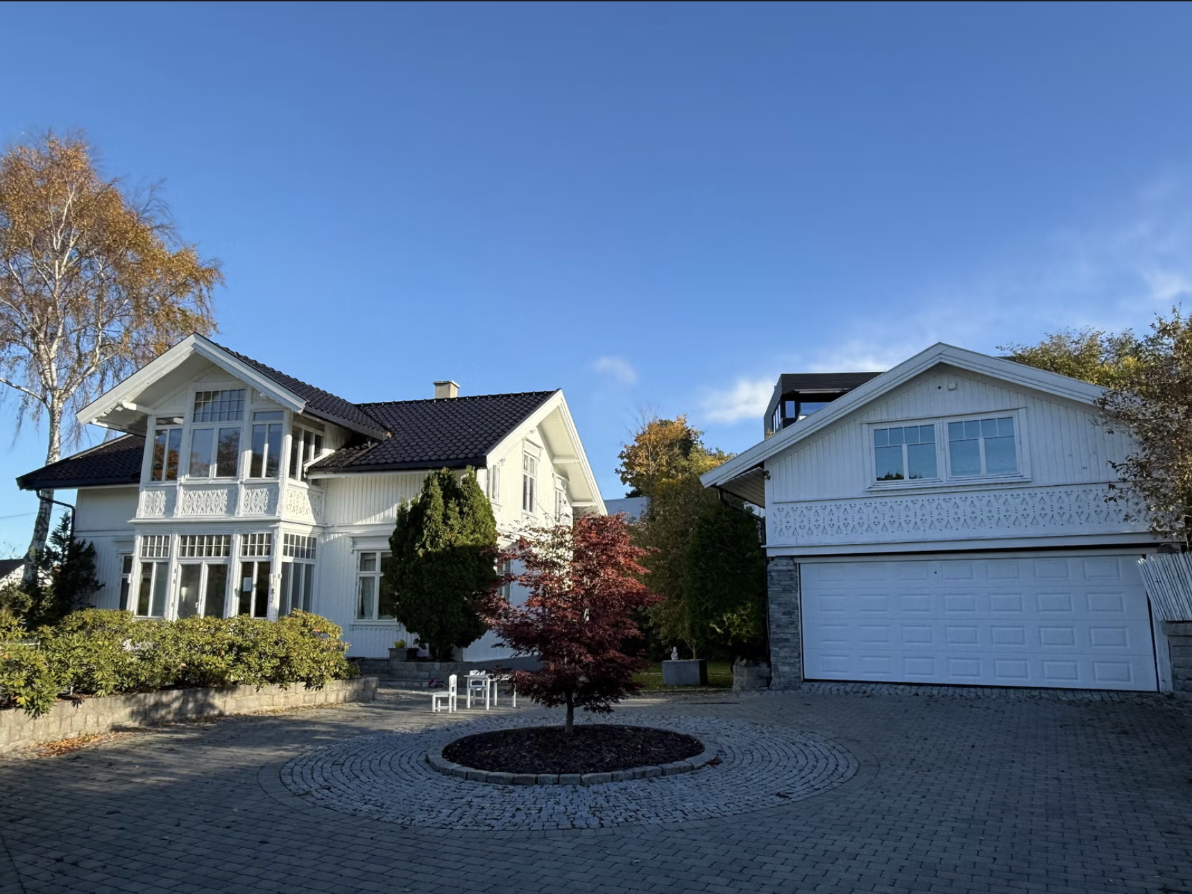 Two white houses with black roofs, surrounded by trees and a paved driveway with a small red tree in the center.