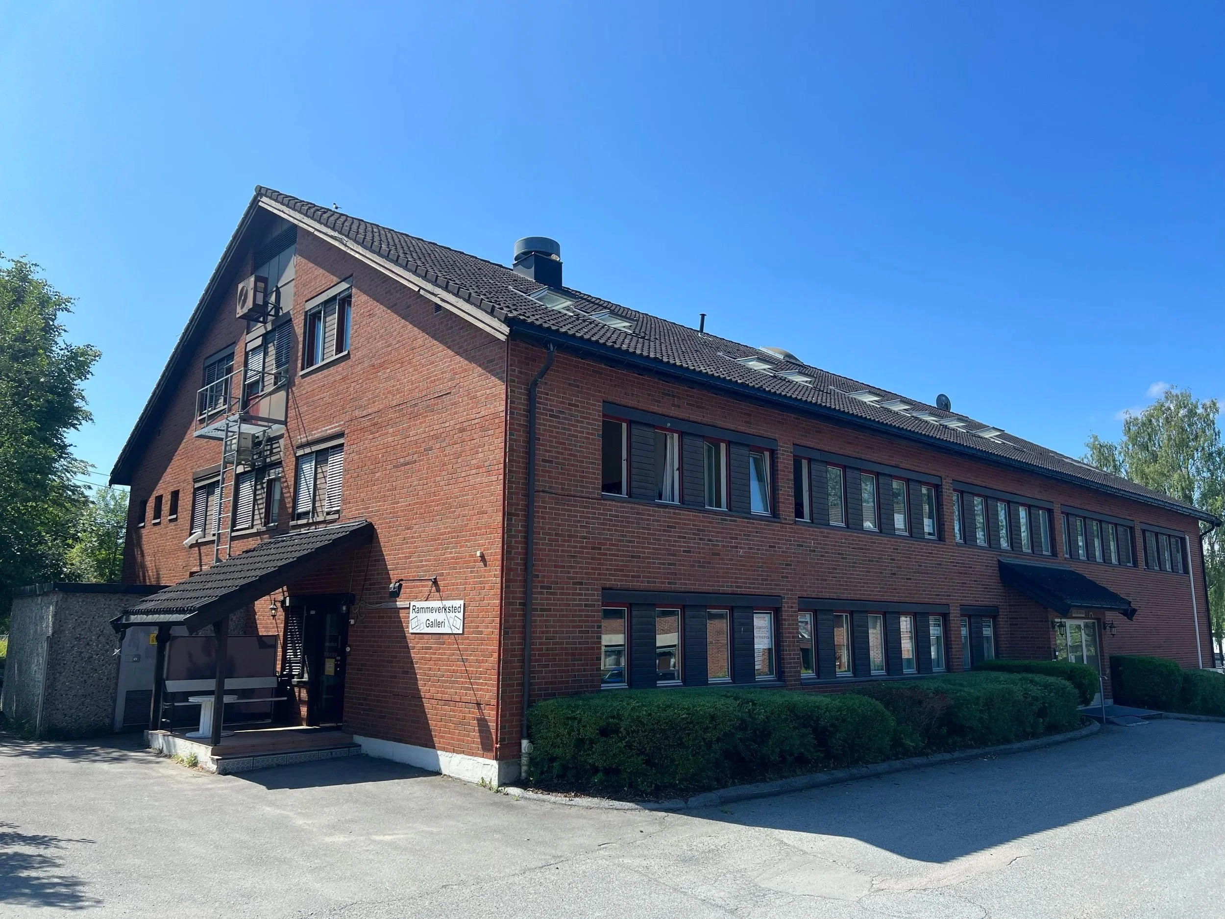 Red brick building with multiple windows and a black tiled roof, situated on a clear, sunny day, surrounded by greenery.