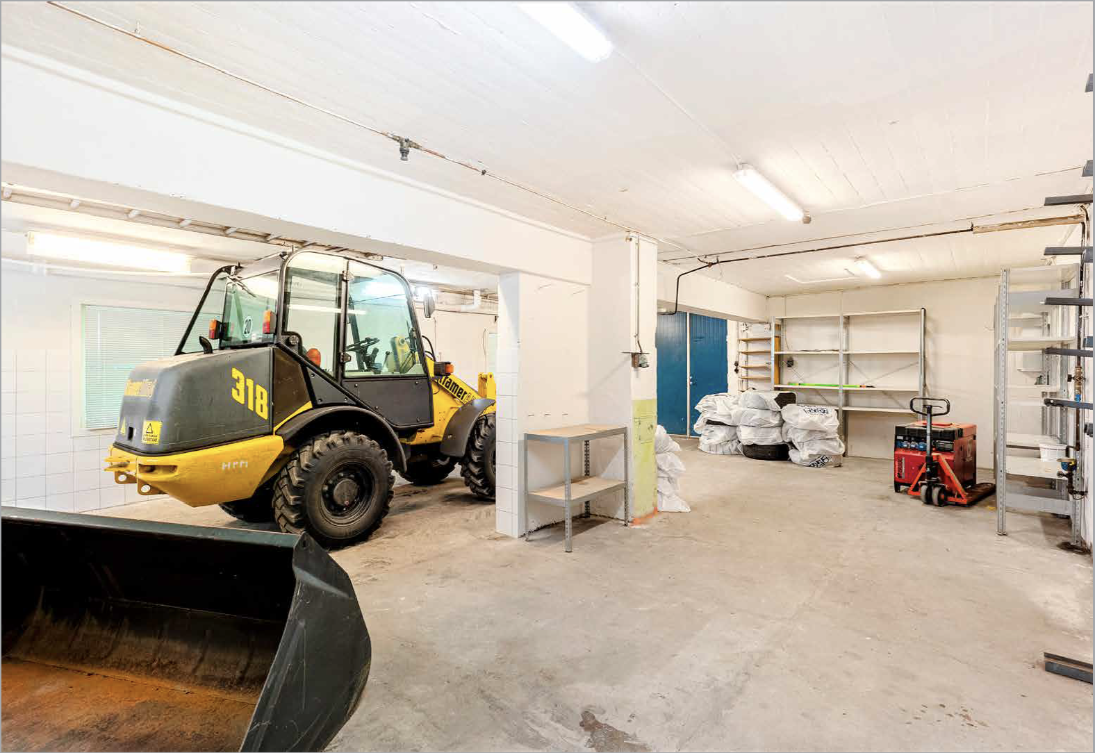 Indoor storage room with a yellow compact loader, metal shelving, bags of materials, and various tools and equipment.