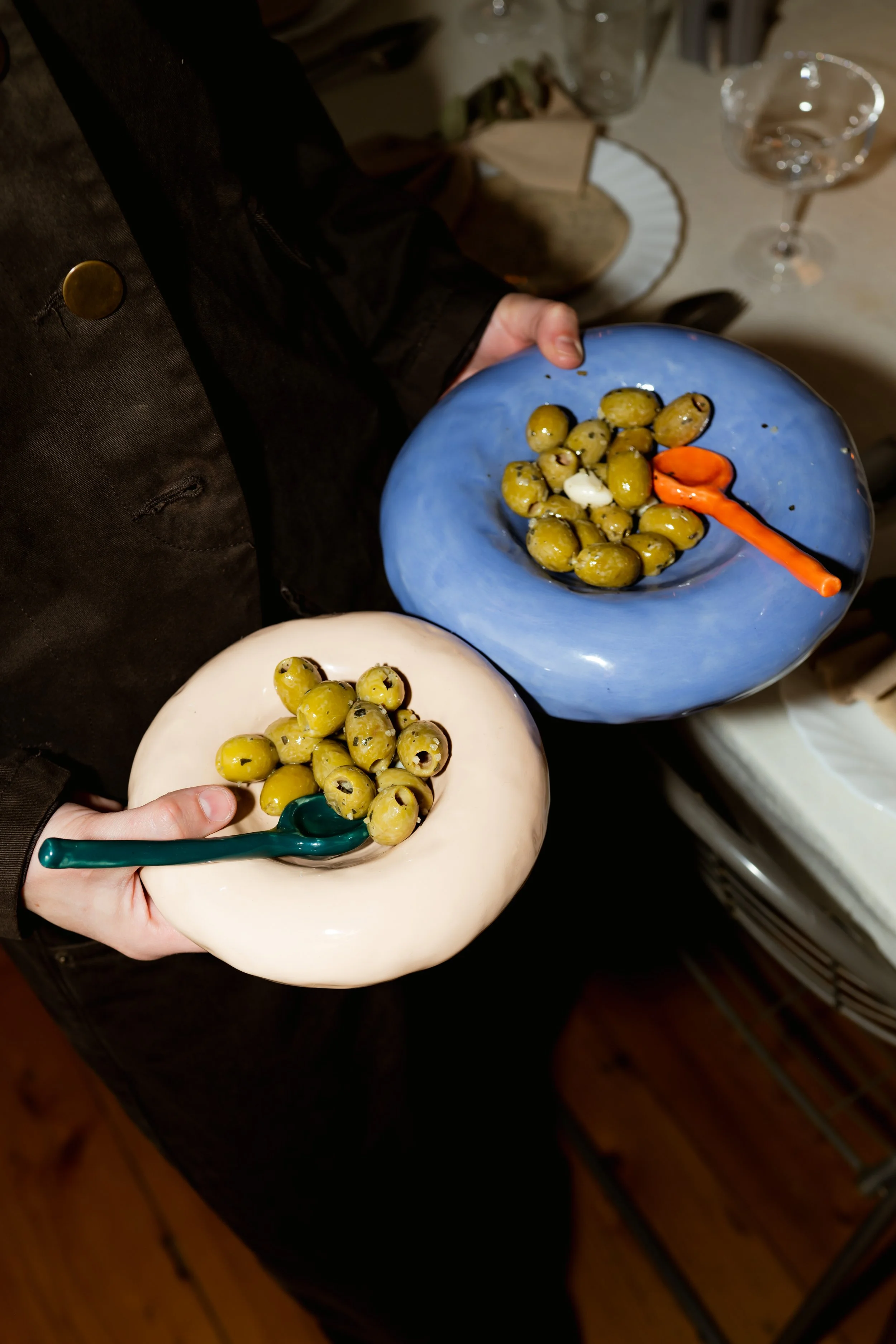 Two people holding ceramic bowls filled with green olives; one bowl is white with a teal spoon, and the other blue with an orange spoon. Den Haag The Hague. Keramiek. klei. ceramics. Workshops. Handopbouw. Handbuilding workshops and Events.