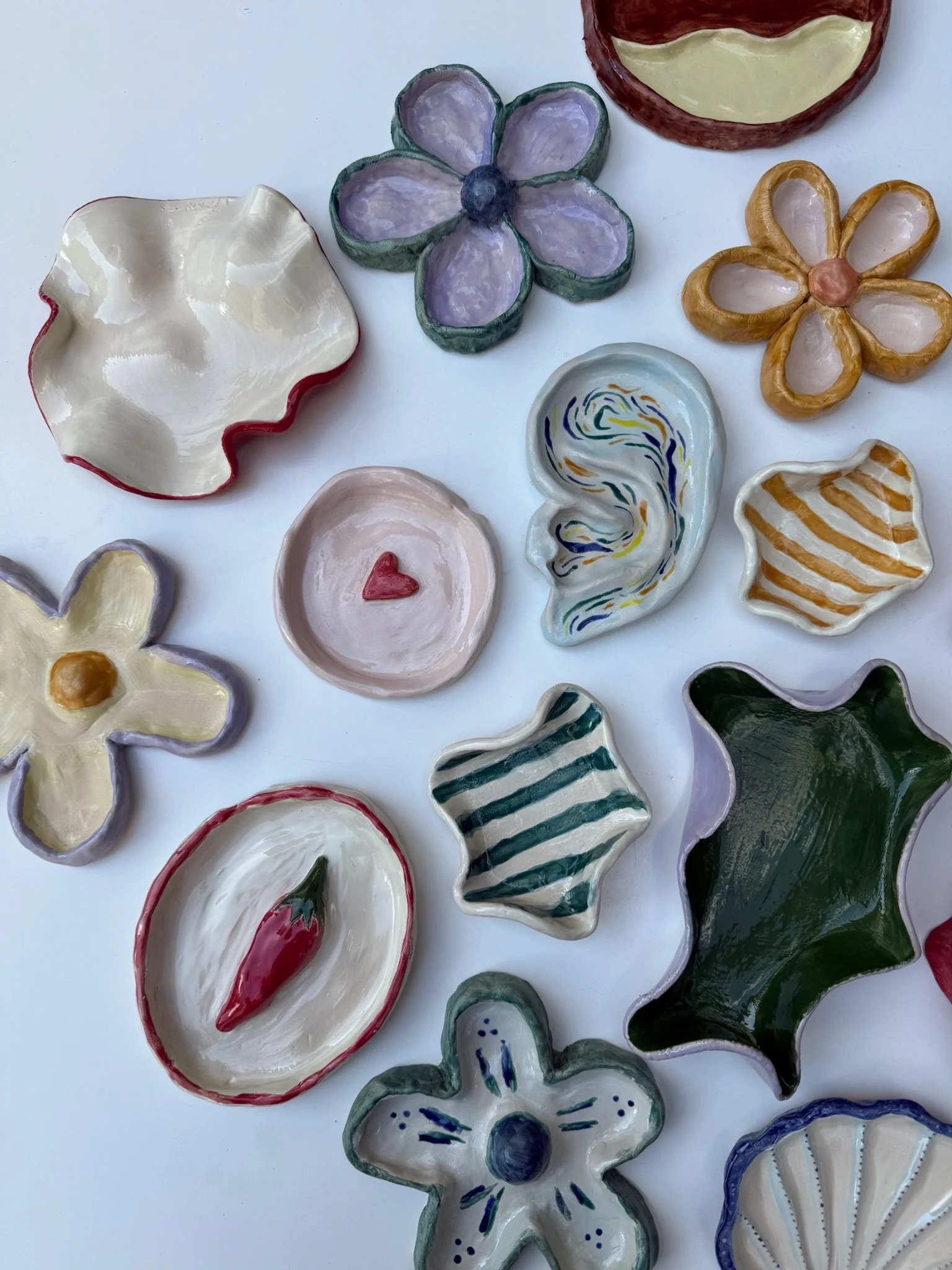 Colorful ceramic dishes shaped like flowers, shells, and leaves, with various patterns and colors, arranged on a white surface. Den Haag The Hague. Keramiek. klei. ceramics. Workshops. Handopbouw. Handbuilding workshops and Events. Groepsuitjes team