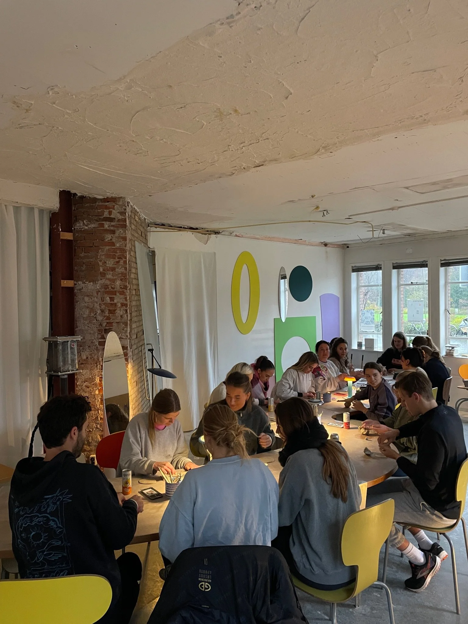 Group of young women and men gathered around a large table in a modern, well-lit room, working on arts and crafts projects.Den Haag The Hague. Keramiek. klei. ceramics. Workshops. Handopbouw. Handbuilding workshops and Events. Groepsuitjes.