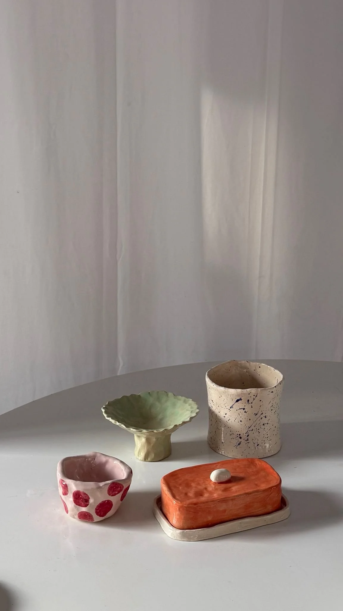 Four handmade ceramic objects on a white table, including a green scalloped bowl, a tall speckled cup, a small bowl with red polka dots, and an orange rectangular dish. Den Haag The Hague. Keramiek. klei. Workshops. Handopbouw. Handbuilding
