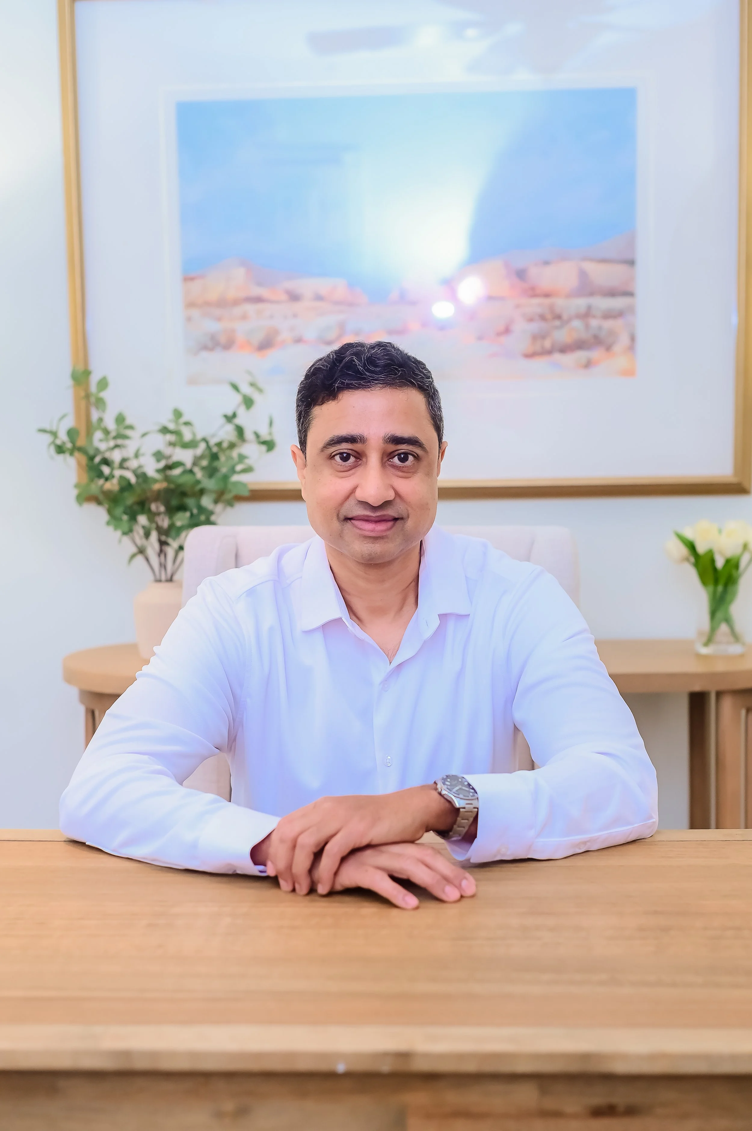 A man in a white dress shirt sitting at a wooden table, with his arms resting on the table, wearing a silver wristwatch, in a room with a large framed desert landscape photograph, a green potted plant, and a vase with white roses behind him.
