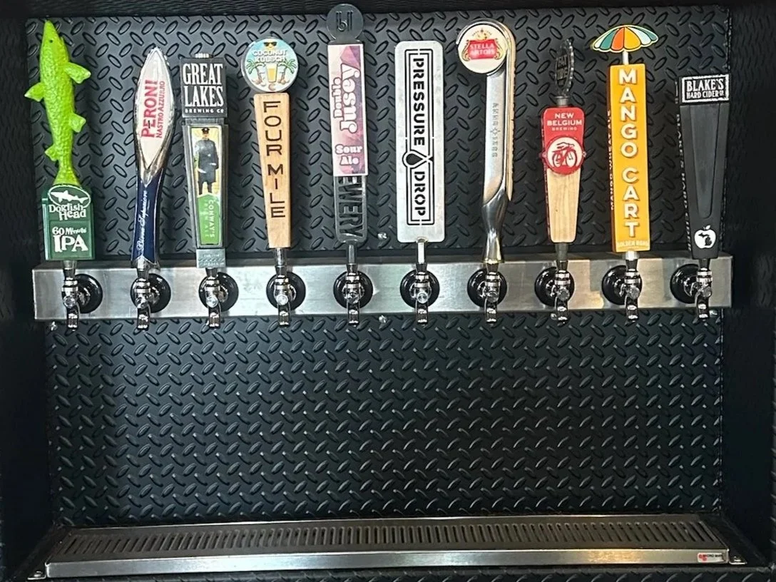 A row of beer tap handles on a black metal background, each labeled with different beer brands and flavors including Peroni, Great Lakes, Sour Ale, Binery, Pressure Drop, Mango Cart, and Blake's Hard Cider.