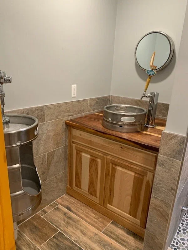 Small restroom with a stainless steel handwashing station, a wooden cabinet with a round metal basin on top, a round mirror above the basin, and beige tile on the lower walls and floor.