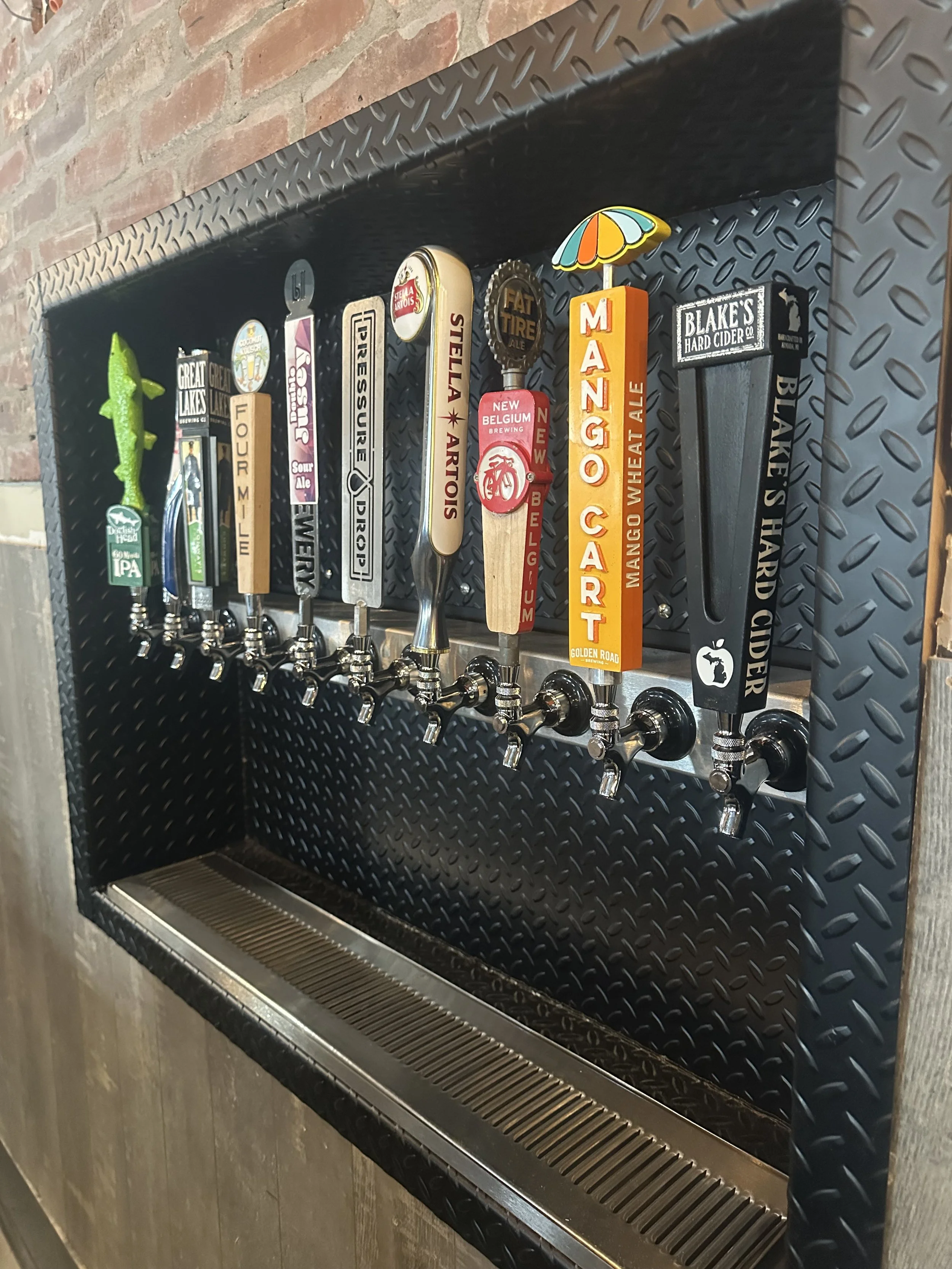 Beer taps with custom handles featuring various brand names and designs, including a green fish, Mango Cart, and Blake's Hard Cider, mounted on a black metal wall with a drip tray below.