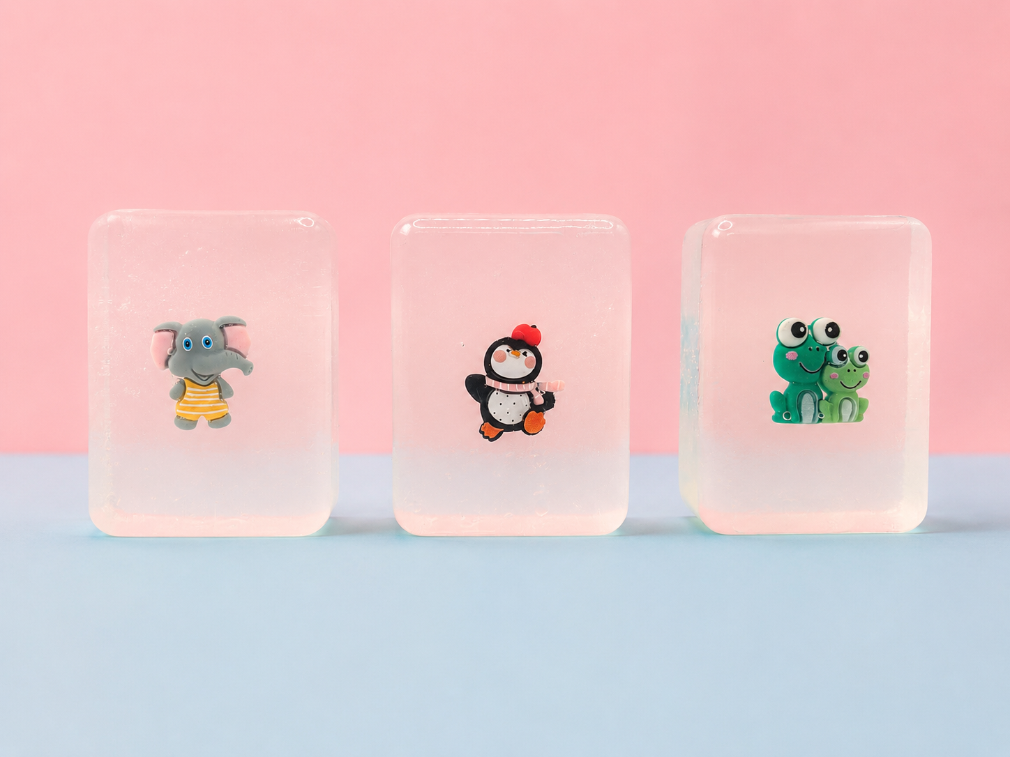 Three transparent hand made soaps, with elephant, pinguin and frog figurines in a pastel blue and pink background