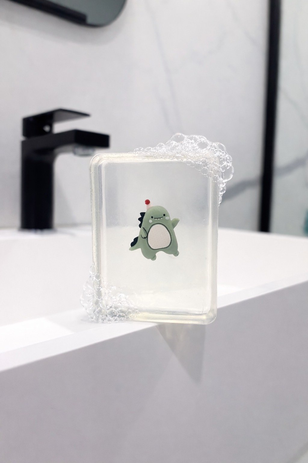 Transparent handmade soap with a collectible dinosaur cute figurine beside a bathroom sink covered in bubbles
