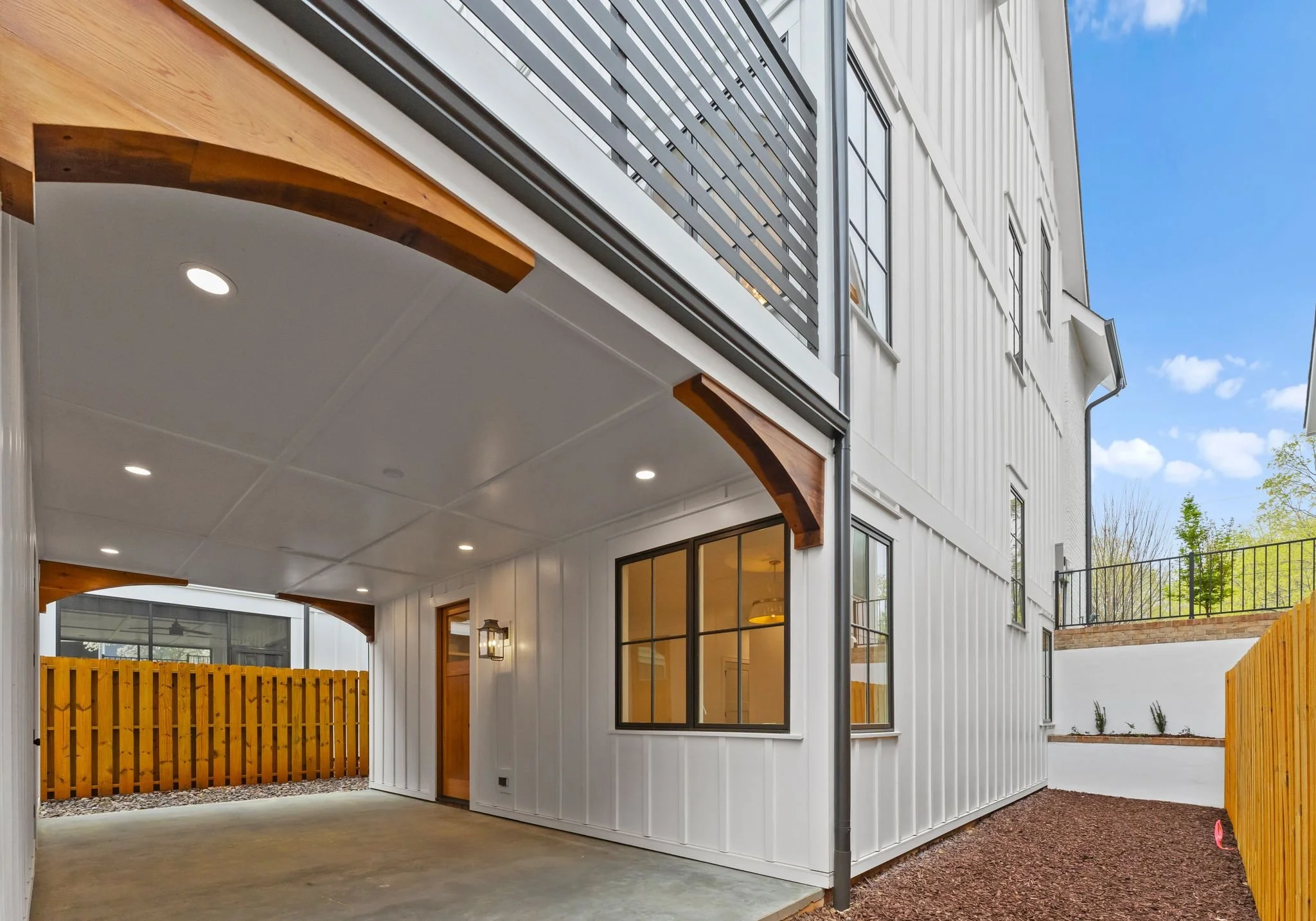 View of a modern white two-story house with black window frames, a wooden fence, a covered patio area with a white ceiling, and a nearby open yard with a gravel pathway and some small plants, under a blue sky with some clouds.
