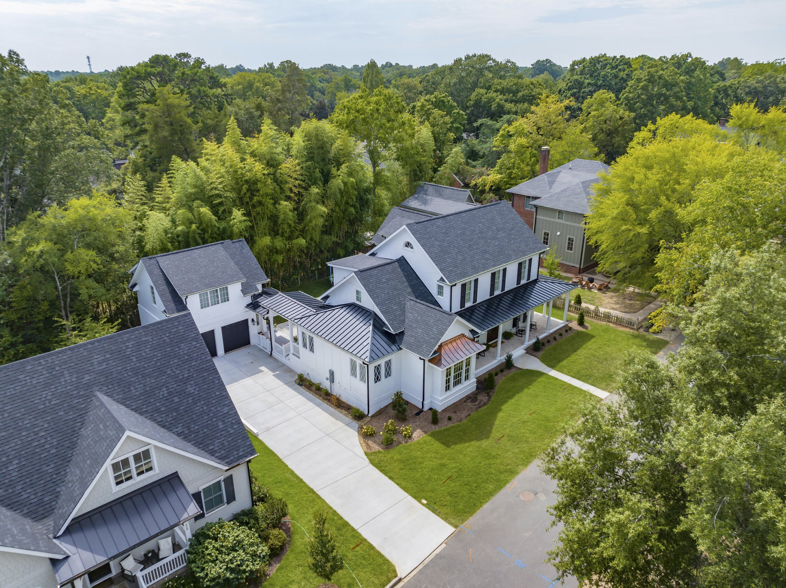 Aerial view of a modern residential neighborhood with a white house featuring a grey roof, surrounded by green trees and a well-maintained lawn, with neighboring houses and a driveway visible.