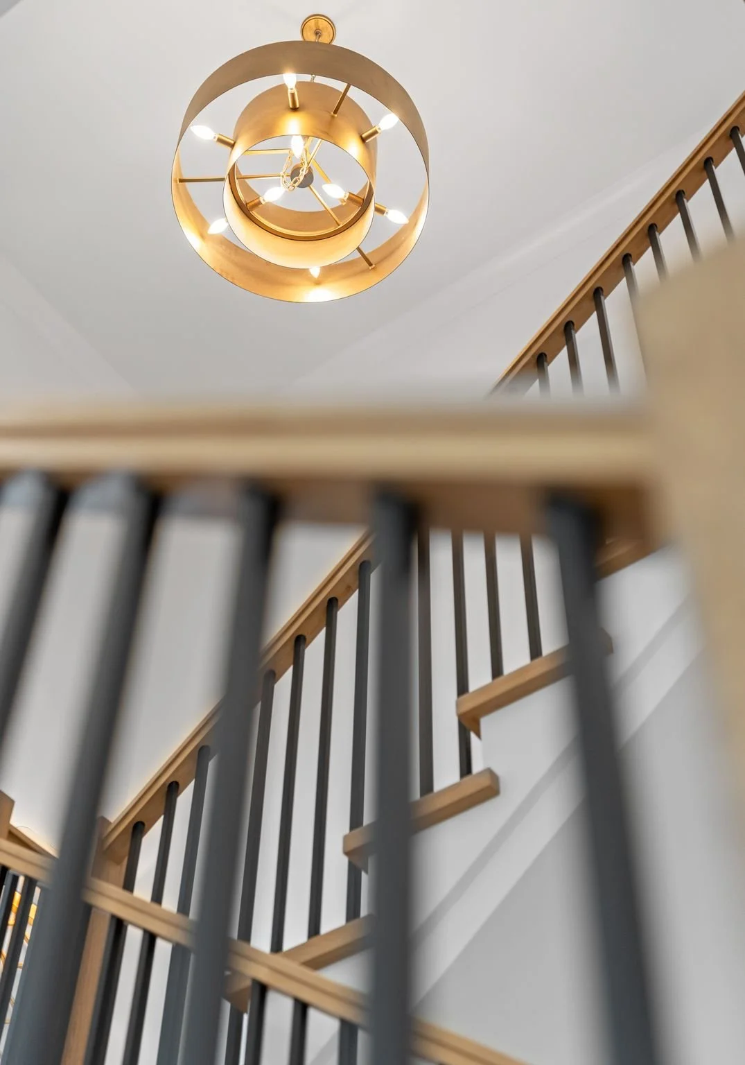 View looking up at a modern gold-colored chandelier hanging from a white ceiling, above a staircase with wooden handrails and black balusters.