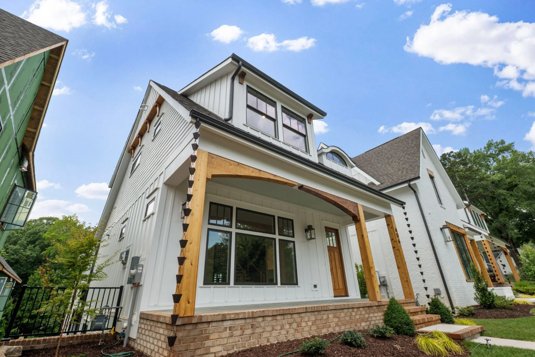 A modern white house with wooden accents, brick foundation, and large windows, situated in a green neighborhood under a blue sky with scattered clouds.