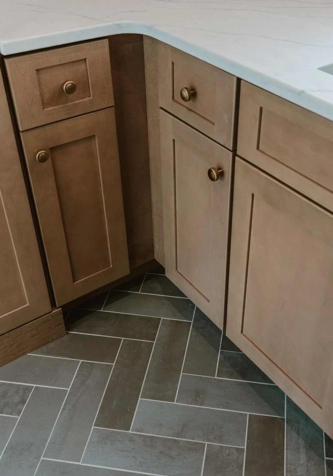 Close-up of a kitchen corner with brown cabinets, gold knobs, and a gray tiled floor.