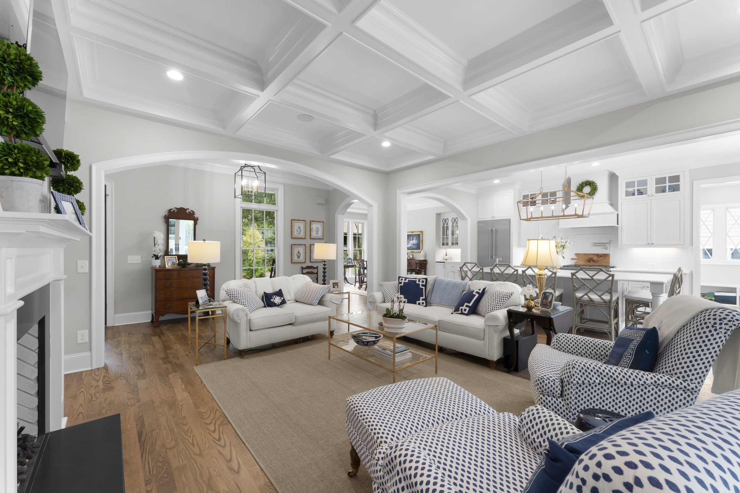 Bright living room with white sofas, patterned armchair, wooden flooring, and large windows, with a view into adjacent room and kitchen.