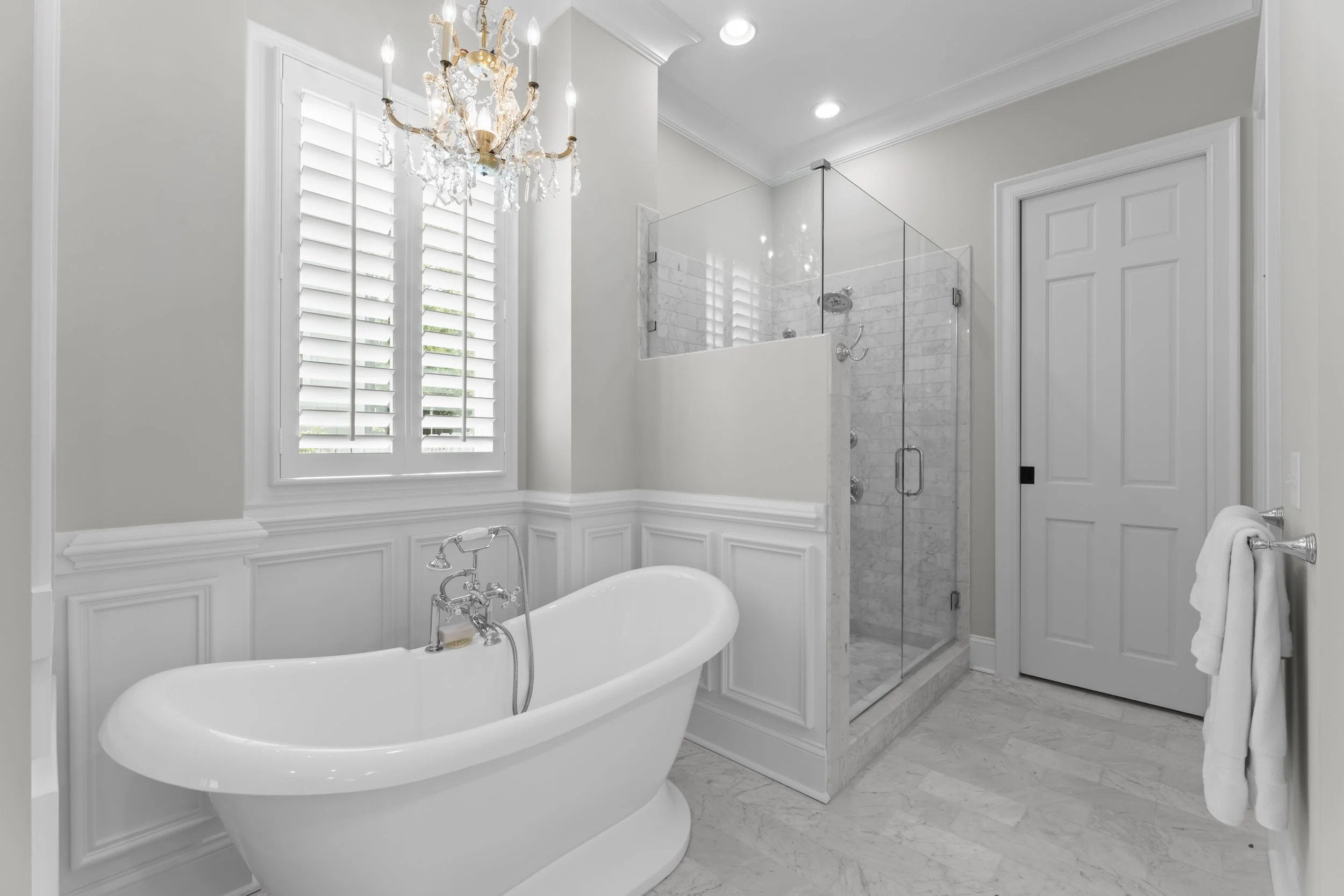 A luxurious bathroom with a freestanding bathtub, a glass-enclosed shower, white paneled walls, and a chandelier hanging from a window with white shutters.
