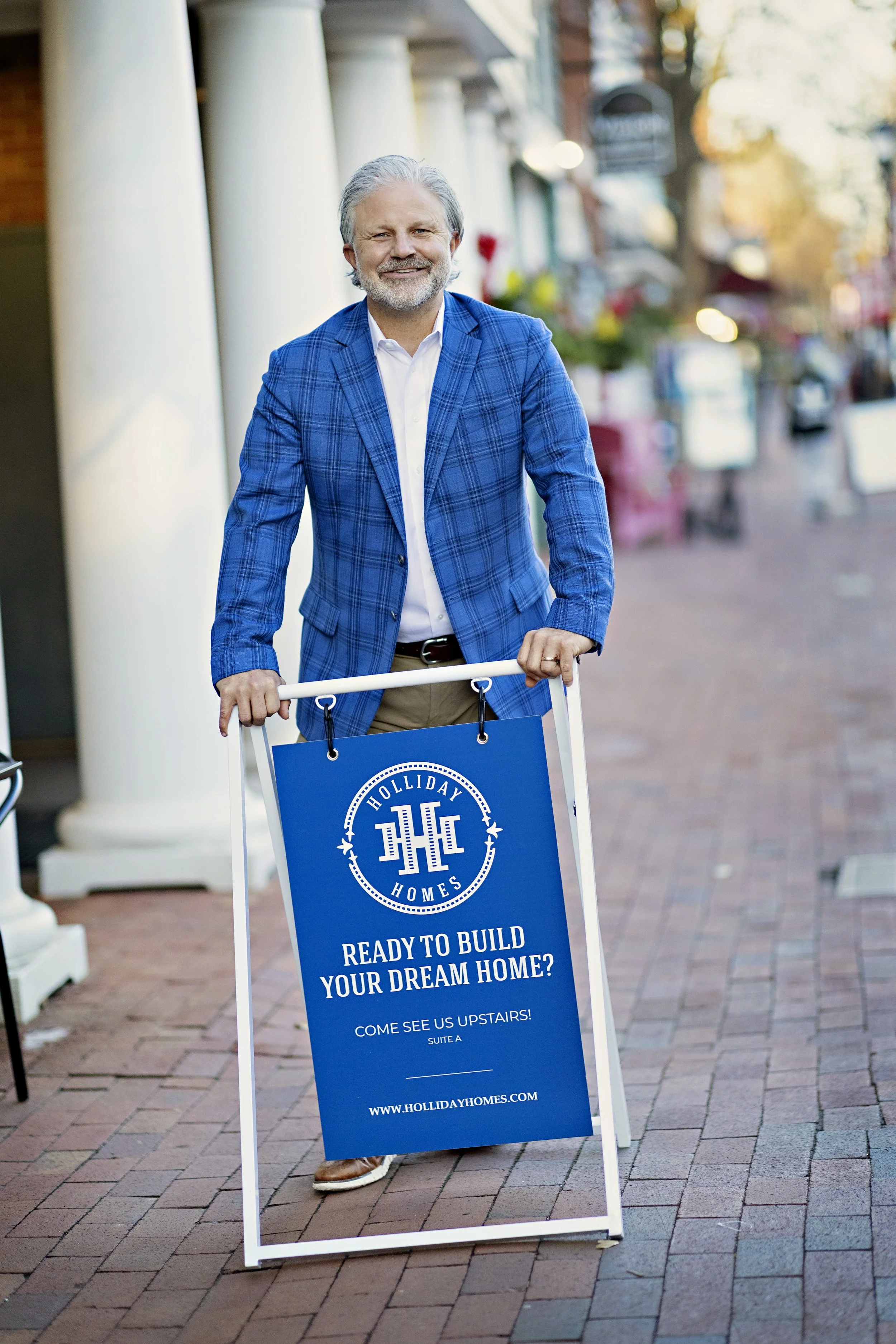A man in a blue plaid blazer holding a sidewalk advertising sign for Holliday Homes, promoting building a dream home, with an outdoor shopping street background.